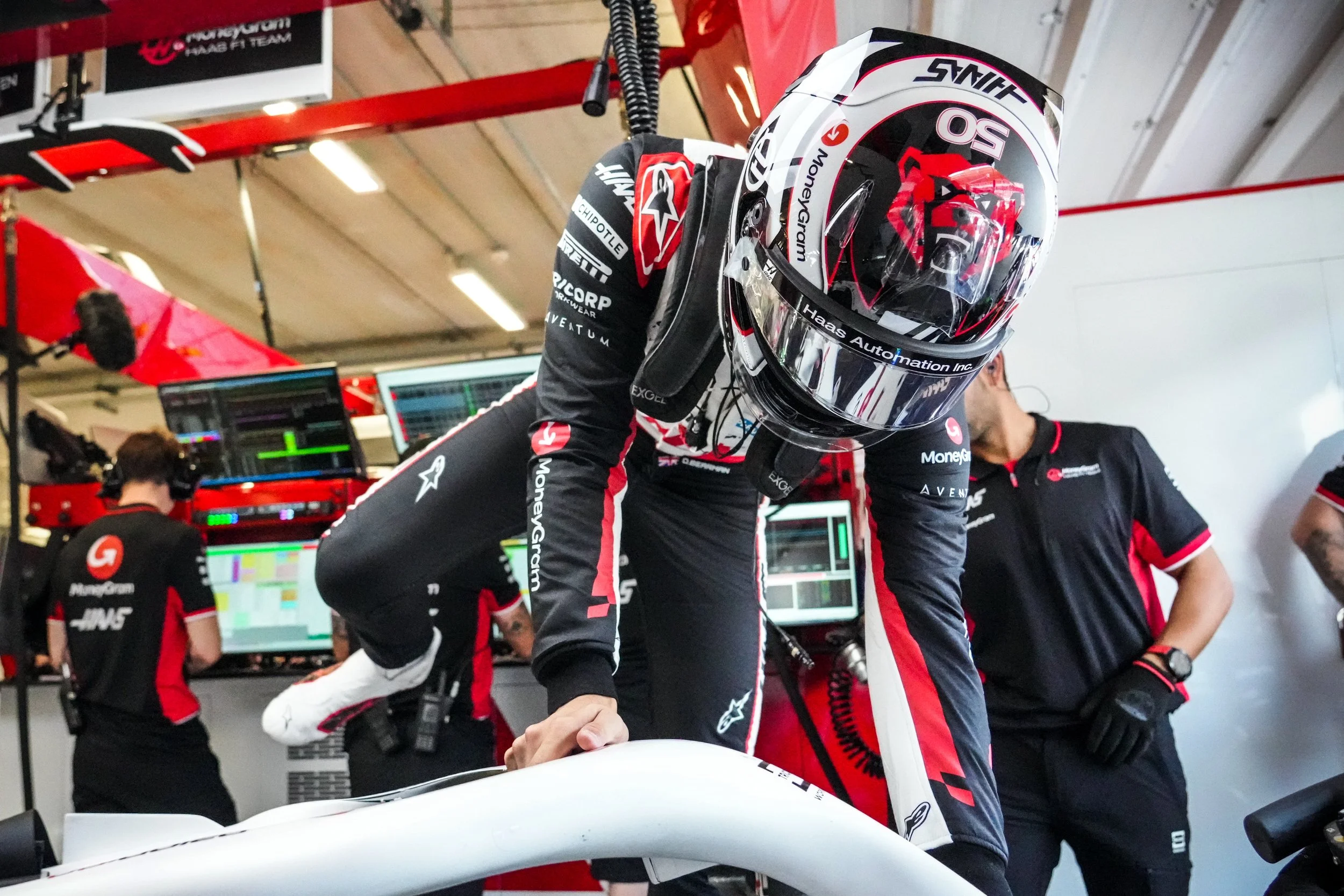 A race car driver in racing gear and helmet leaning over a race car, surrounded by team members in a garage or pit area with monitors and equipment in the background.