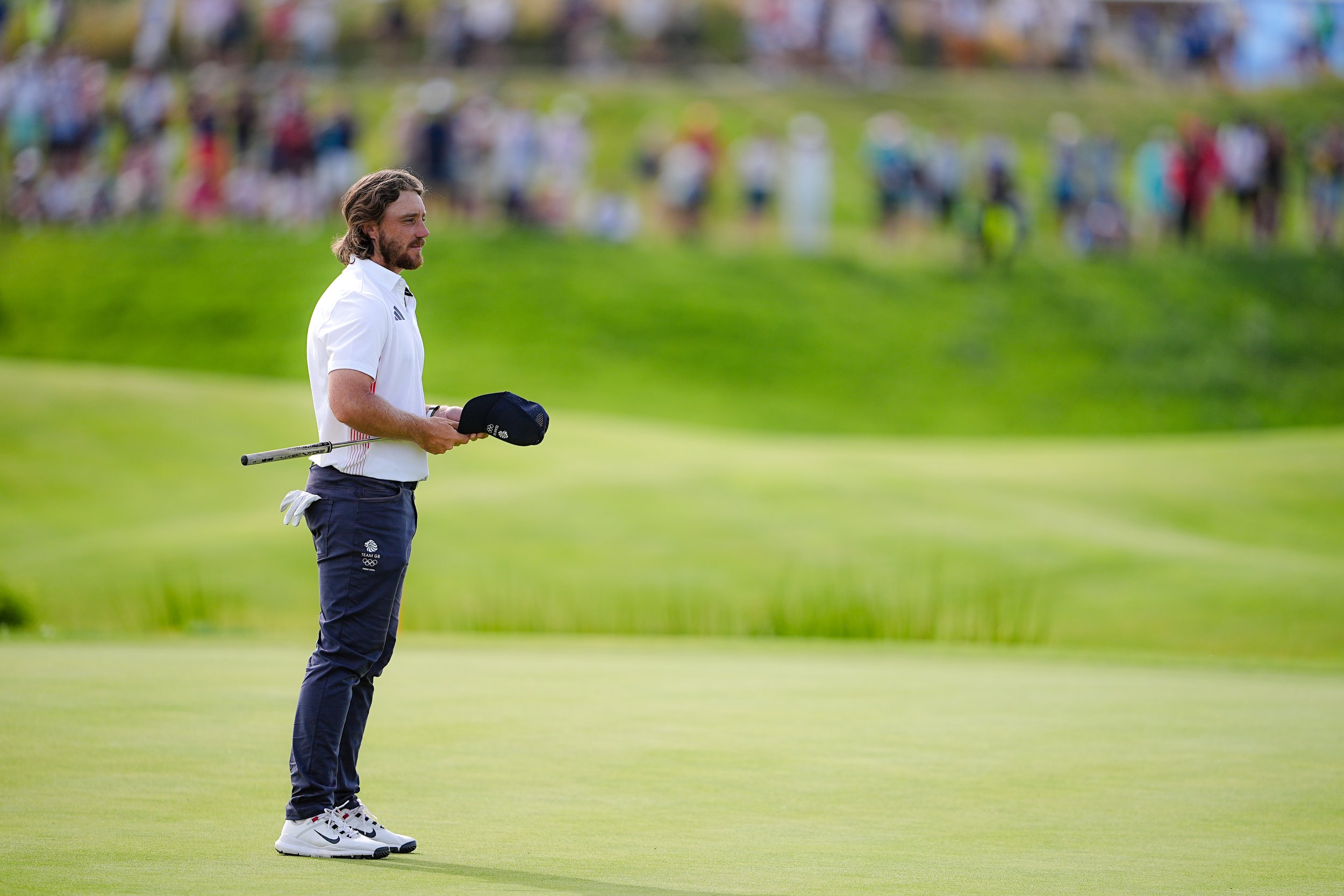 A man with long hair and a beard, dressed in a white shirt and navy pants, stands on a golf course holding a golf cap and a putter. In the background, there is a blurred crowd of spectators.