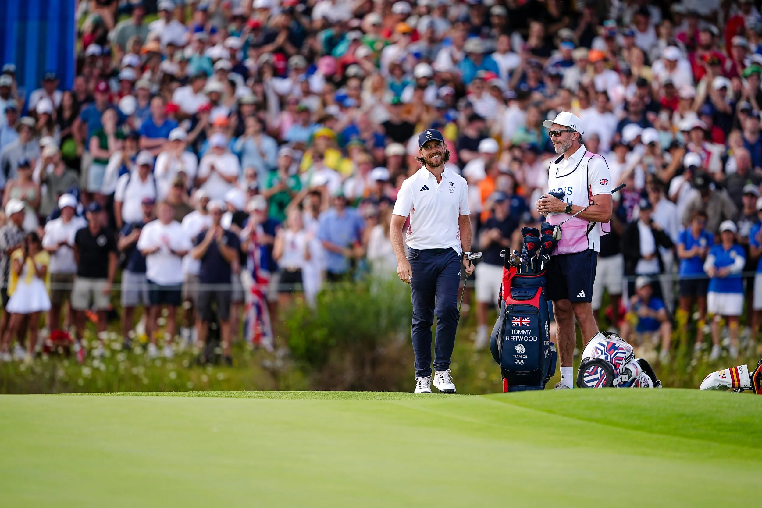 Two male golfers with golf bags on a lush green golf course, with a large crowd of spectators in the background.