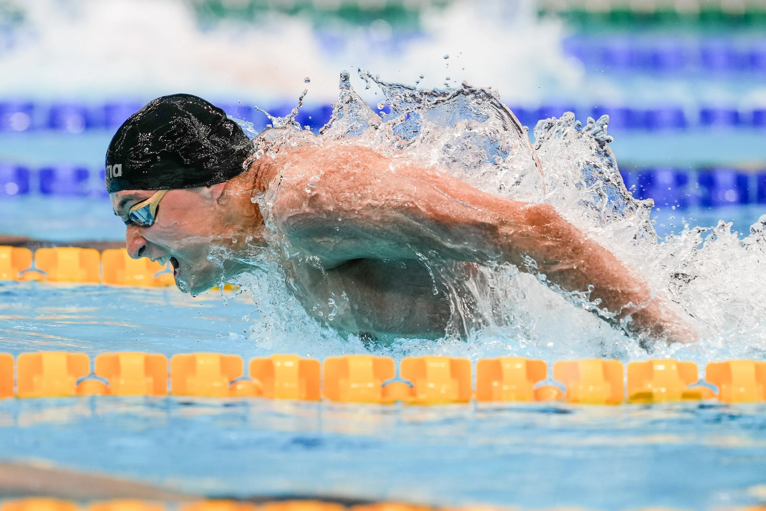 Swimmer in a black swim cap and goggles competing in a swimming race, performing the butterfly stroke in a pool with orange lane dividers.