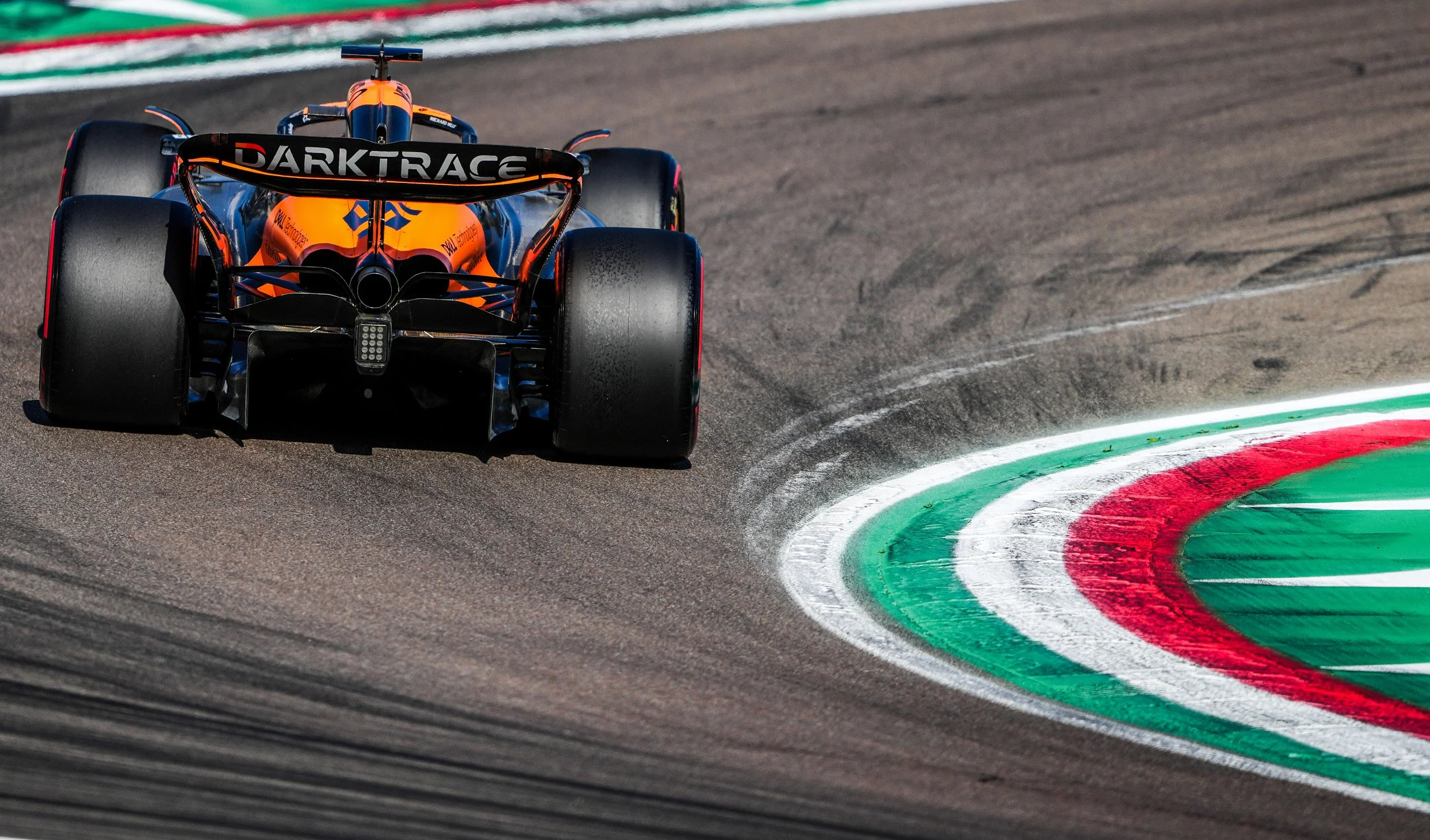 Formula 1 race car on a racetrack, approaching a turn with painted green, white, and red curb markings, viewed from the rear.
