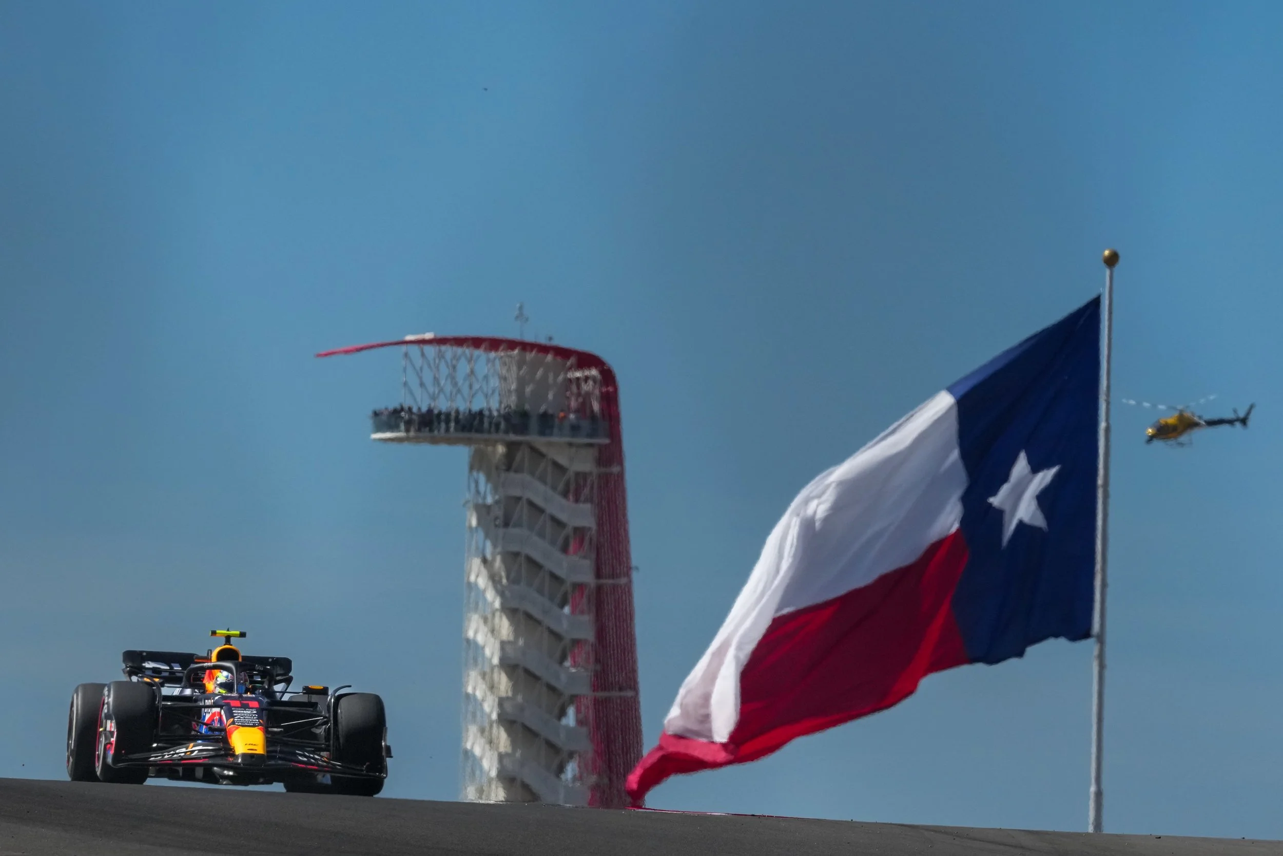 A red bull racing Formula 1 car on a racetrack, with the Texas flag flying on a pole, a yellow helicopter in the sky, and a tall observation tower in the background against a blue sky.