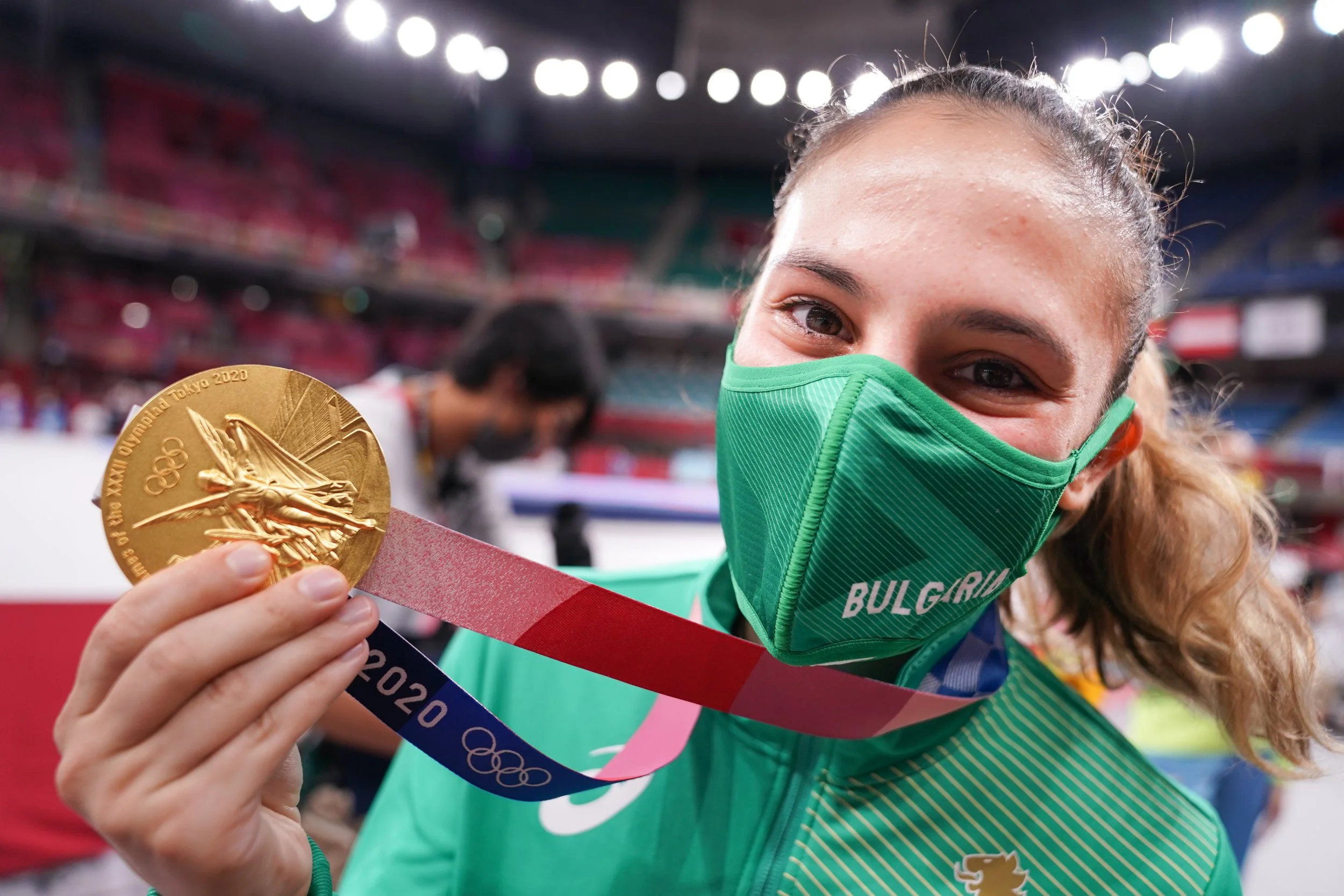 A female athlete from Bulgaria wearing a green face mask and green sports jacket, holding a gold Olympic medal with a pink, red, and blue ribbon at the Tokyo 2020 Olympics. She is smiling and standing in an indoor stadium.