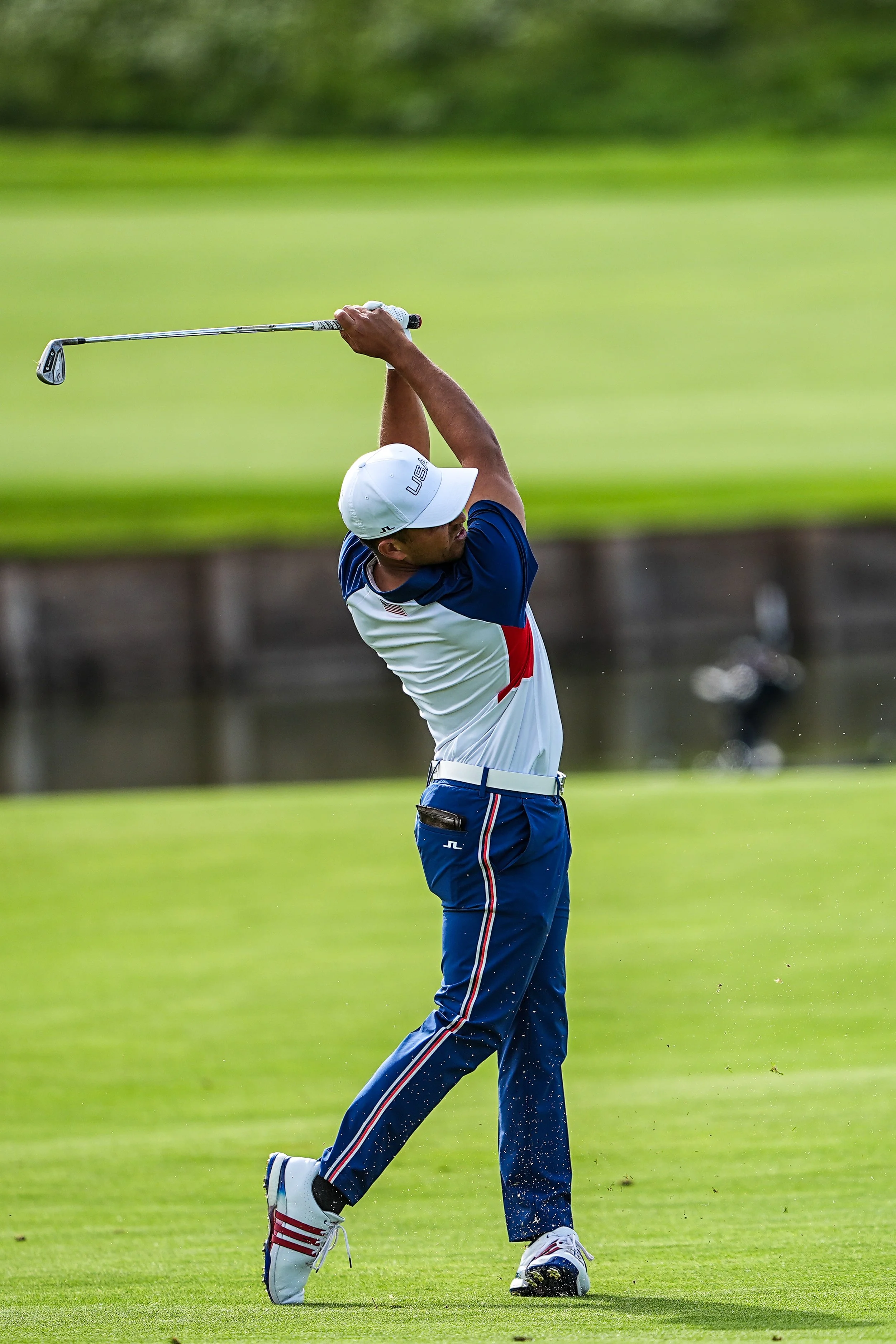 A male golfer in a white cap, white shirt with blue and red accents, and blue pants is mid-swing on a golf course.