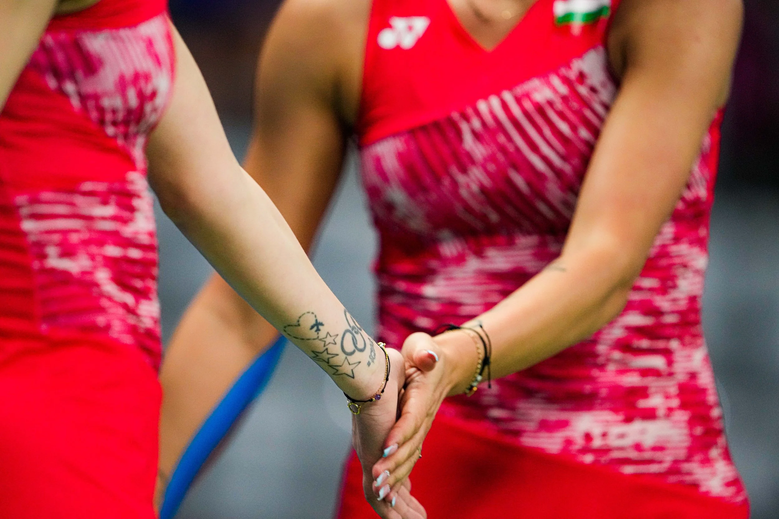 Two athletes in red sports uniforms are holding hands, with one person's arm visible showing tattoos and bracelets.