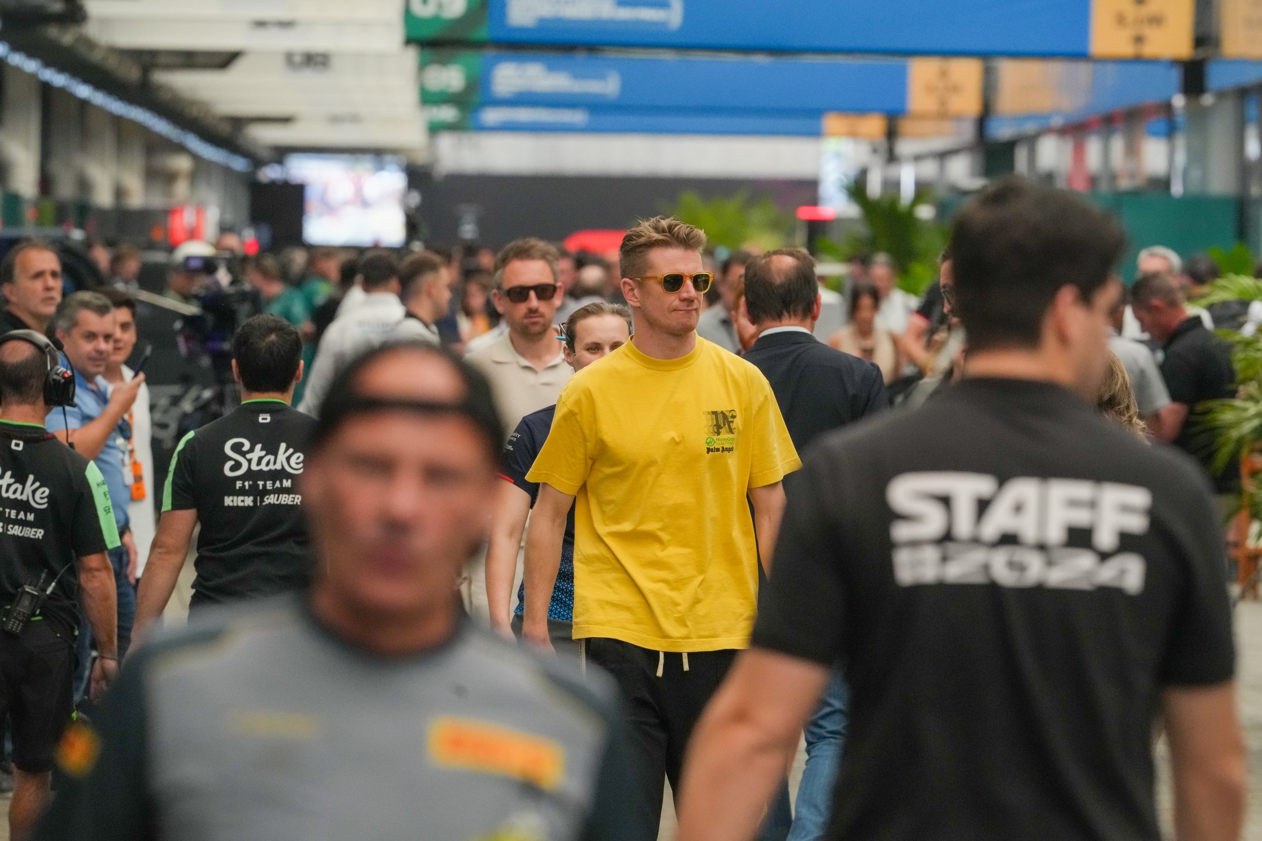 Crowd of people at a racing event, including a man in a yellow shirt and sunglasses surrounded by staff and spectators.