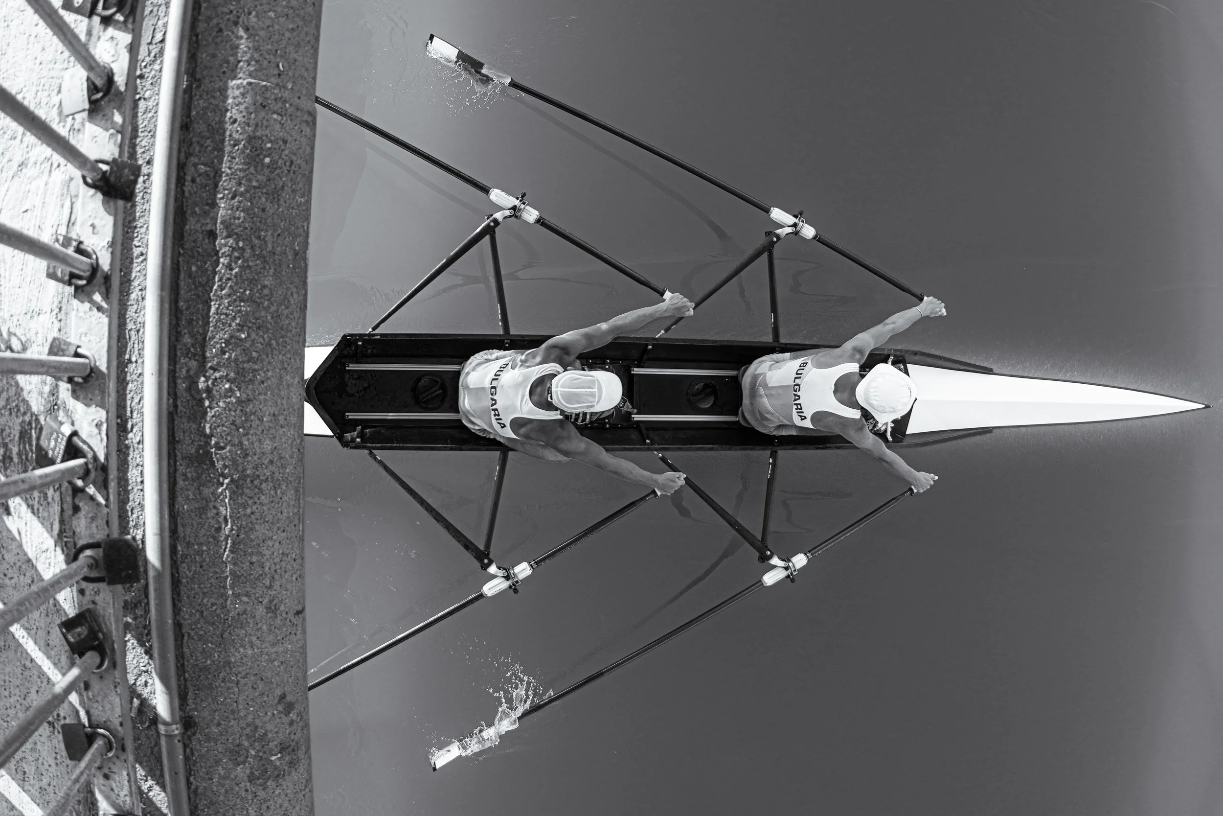 Two rowers in a shell boat with 'Bulgaria' written on their uniforms, seen from above, rowing on a body of water.