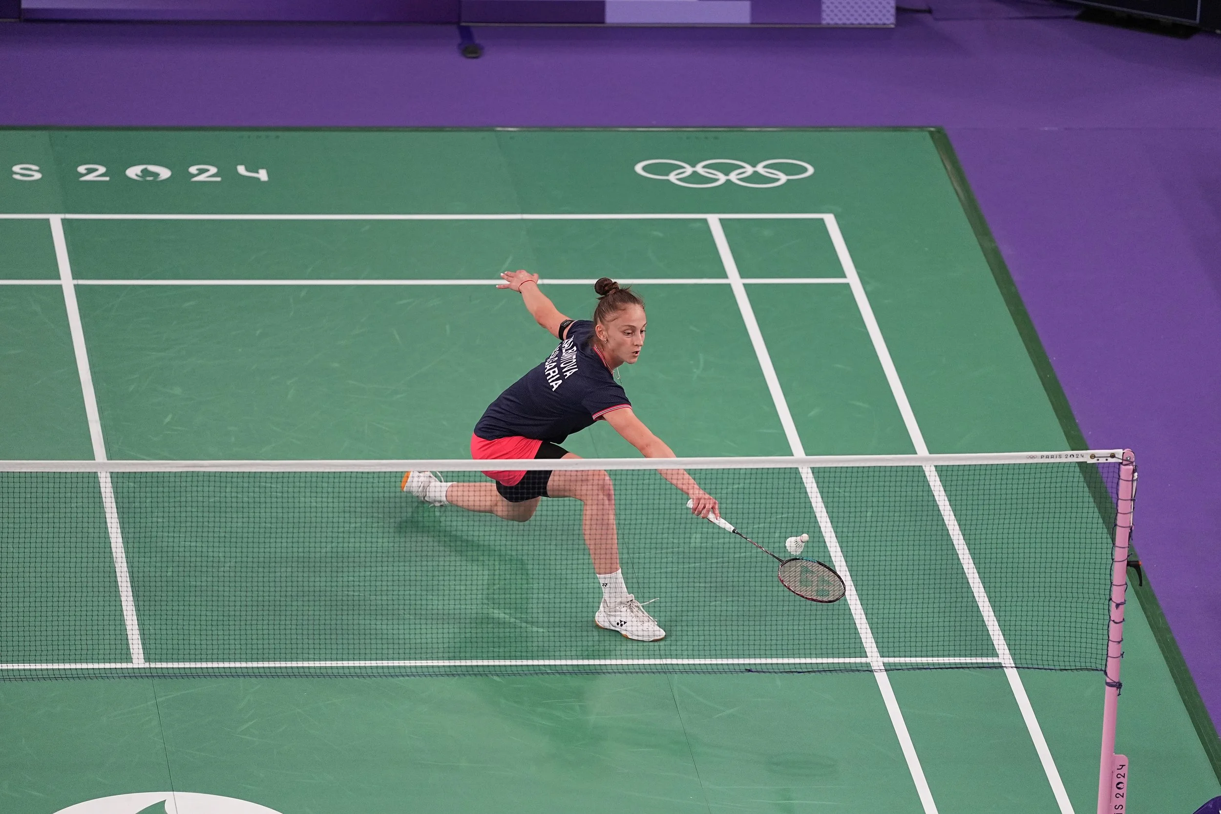 A female badminton player in a black shirt and red shorts lunges on a green badminton court with white lines, hitting a shuttlecock with her racket. The court features the Olympic rings logo.