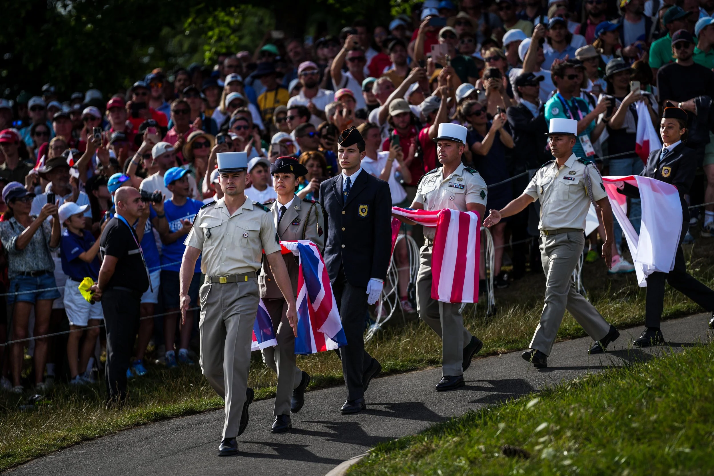Military personnel carrying flags during a formal ceremony, with a large crowd of spectators watching.