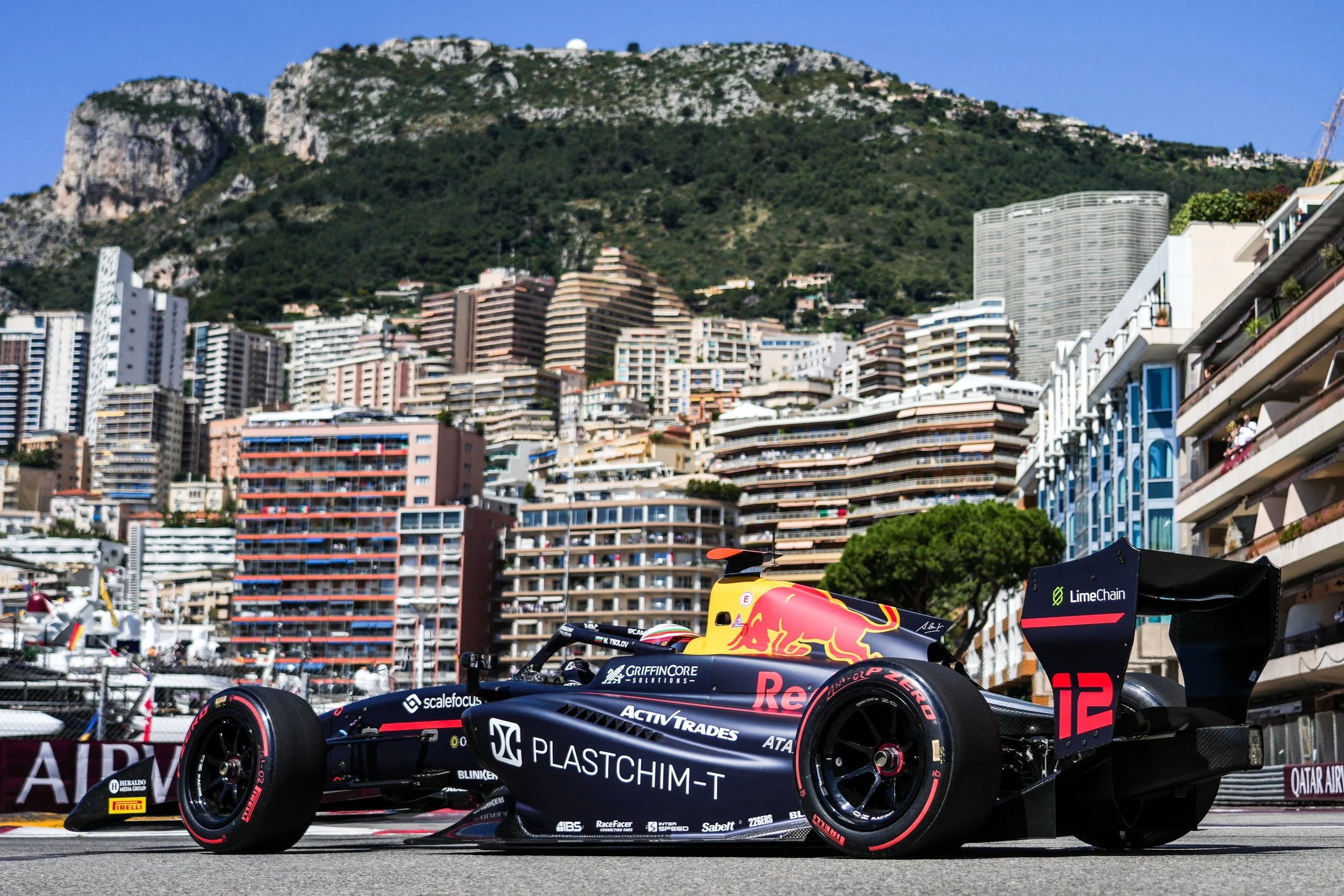 A dark blue race car with Red Bull and various sponsor logos parked on a street with high-rise buildings and a mountain in the background.
