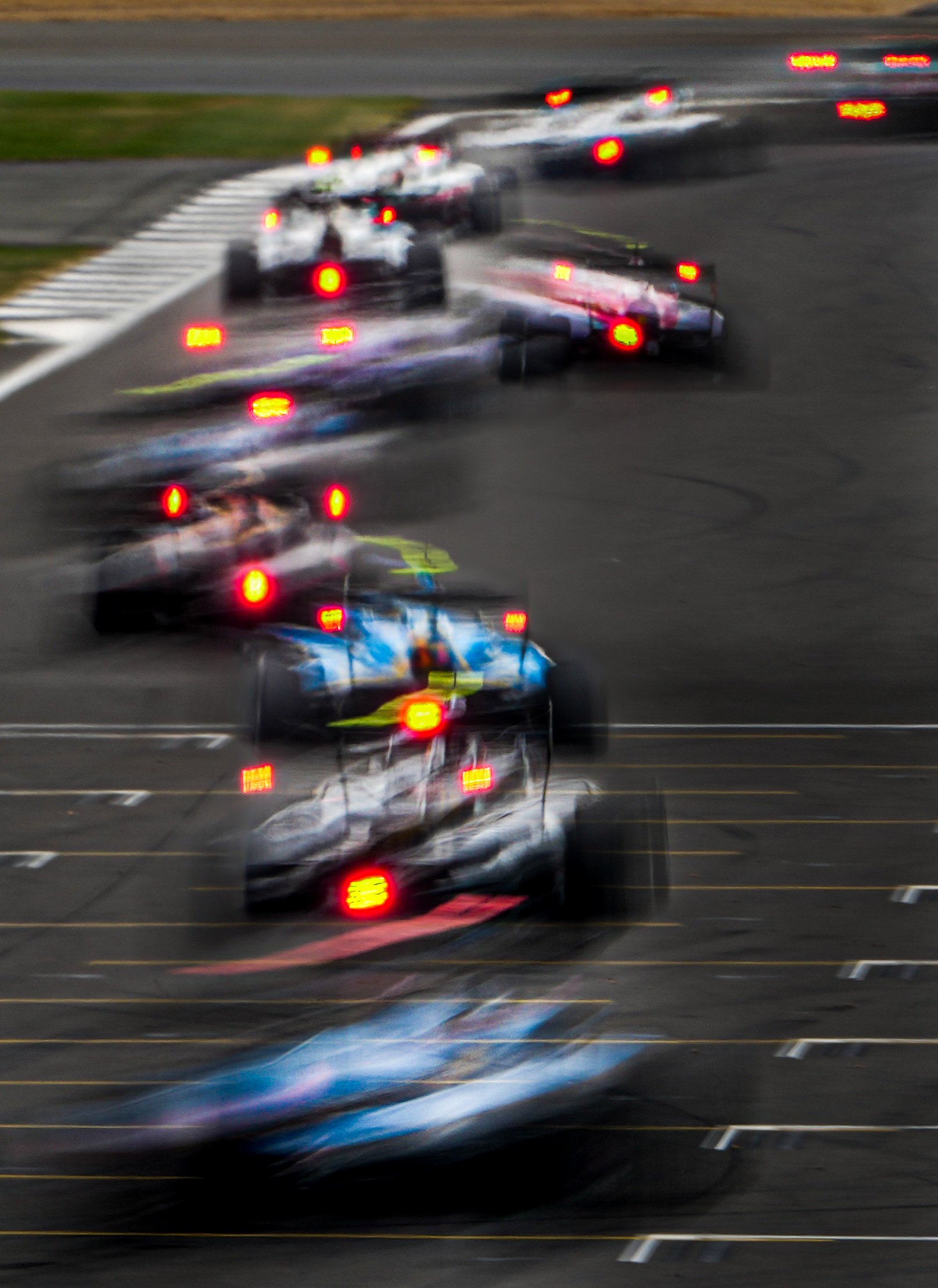 Blurred photo of race cars on a track with many red taillights, indicating a race start or finish.