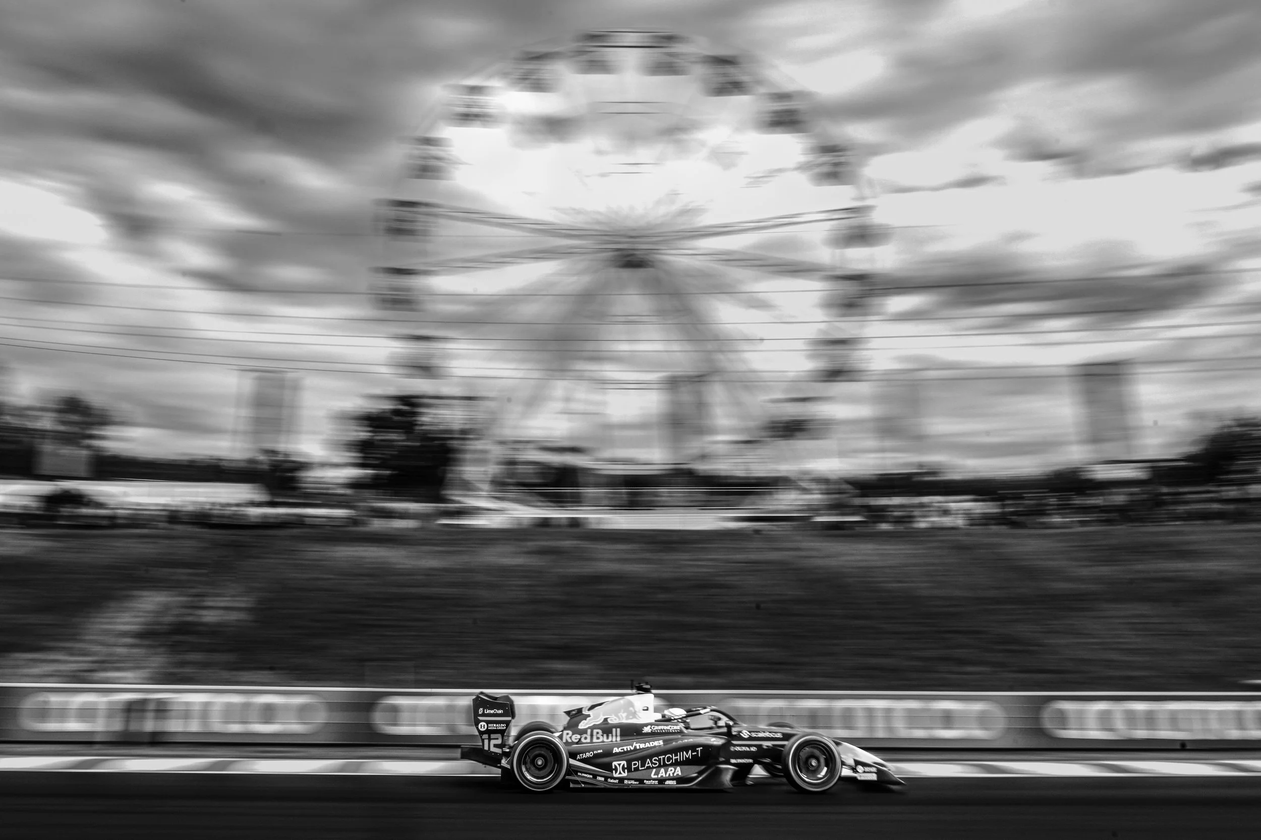 A black and white photograph of a Formula racing car moving at high speed on a track in front of a blurred amusement park Ferris wheel and cloudy sky.