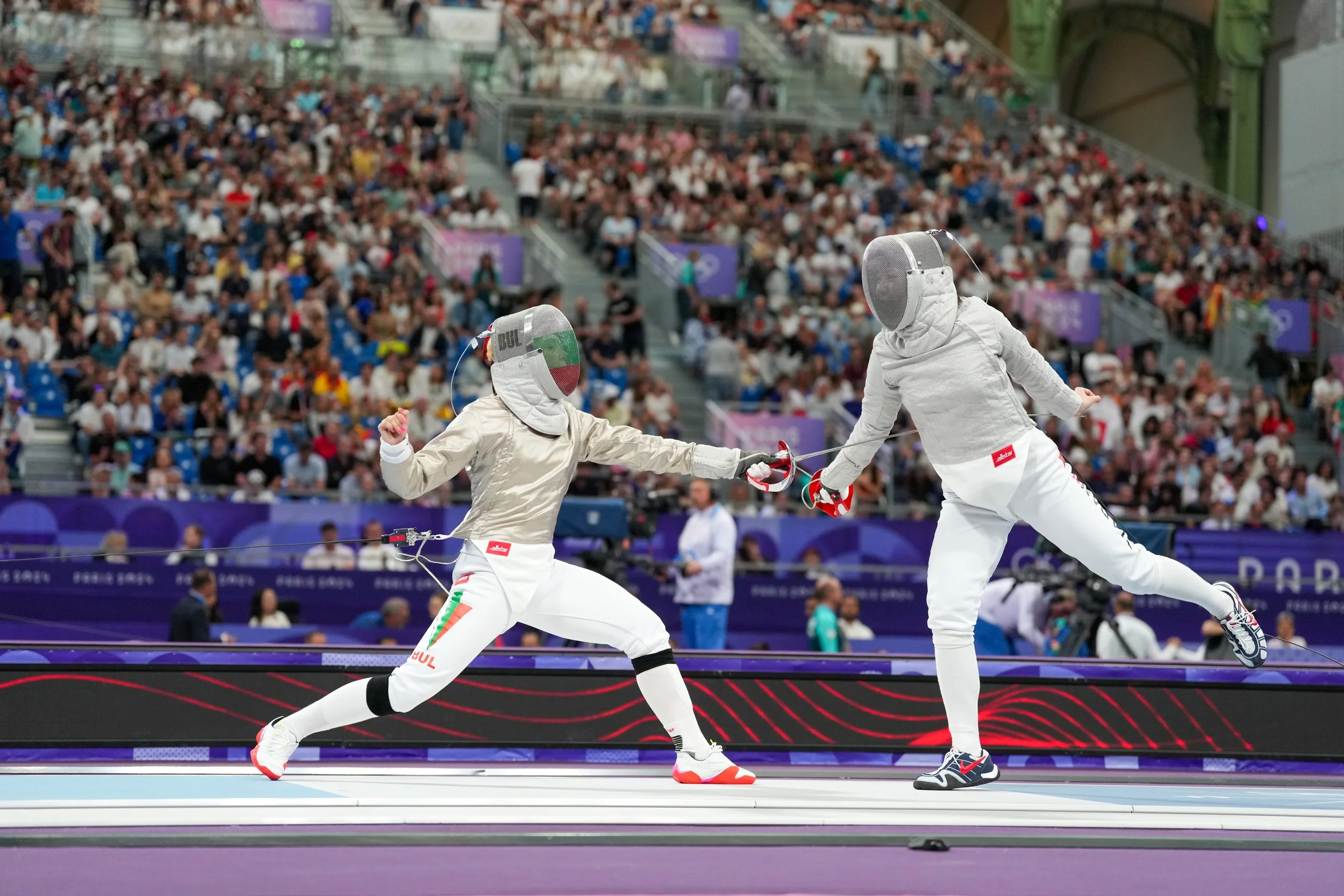 Two fencers in white uniforms and face masks fencing on a purple strip, with a large crowd of spectators watching in the background.