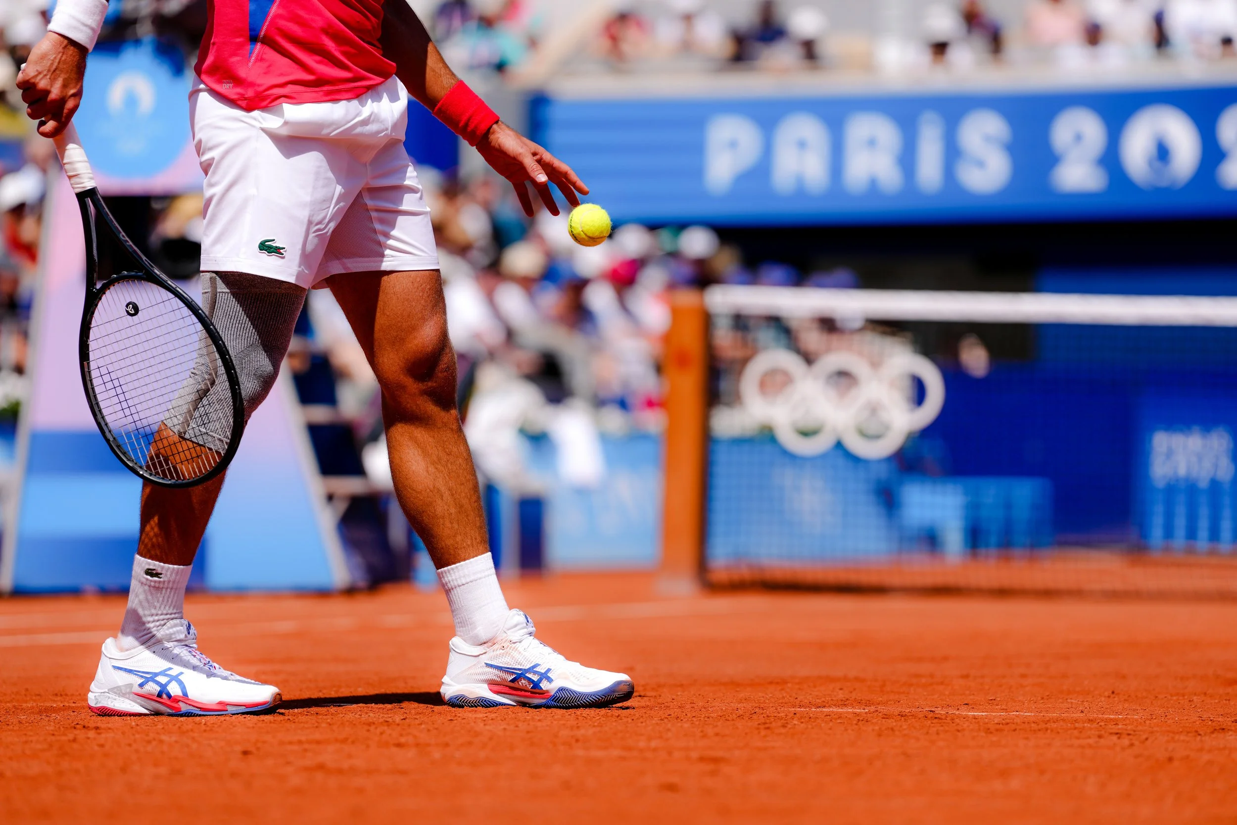 Tennis player on clay court holding tennis ball with tennis racket, wearing white shorts, white shoes with blue and red accents, and a red wristband, during day time at the Paris 2024 Olympics.