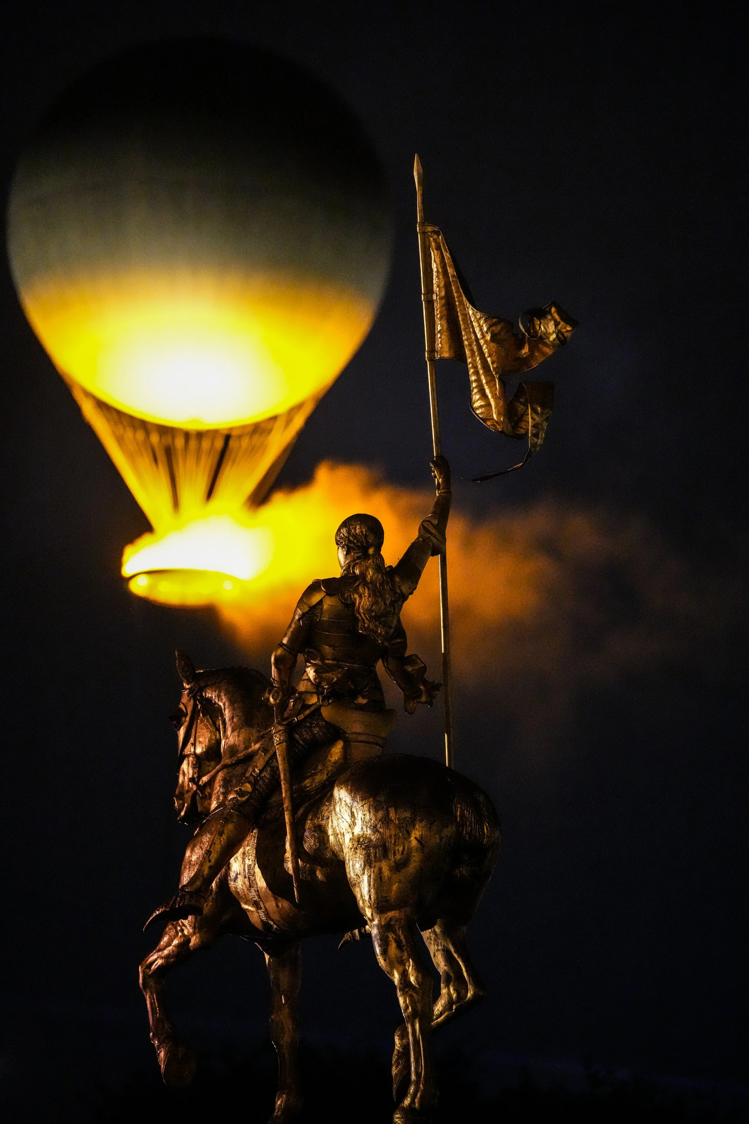 Bronze statue of a mounted soldier holding a flag, with a large, illuminated hot air balloon in the night sky in the background.