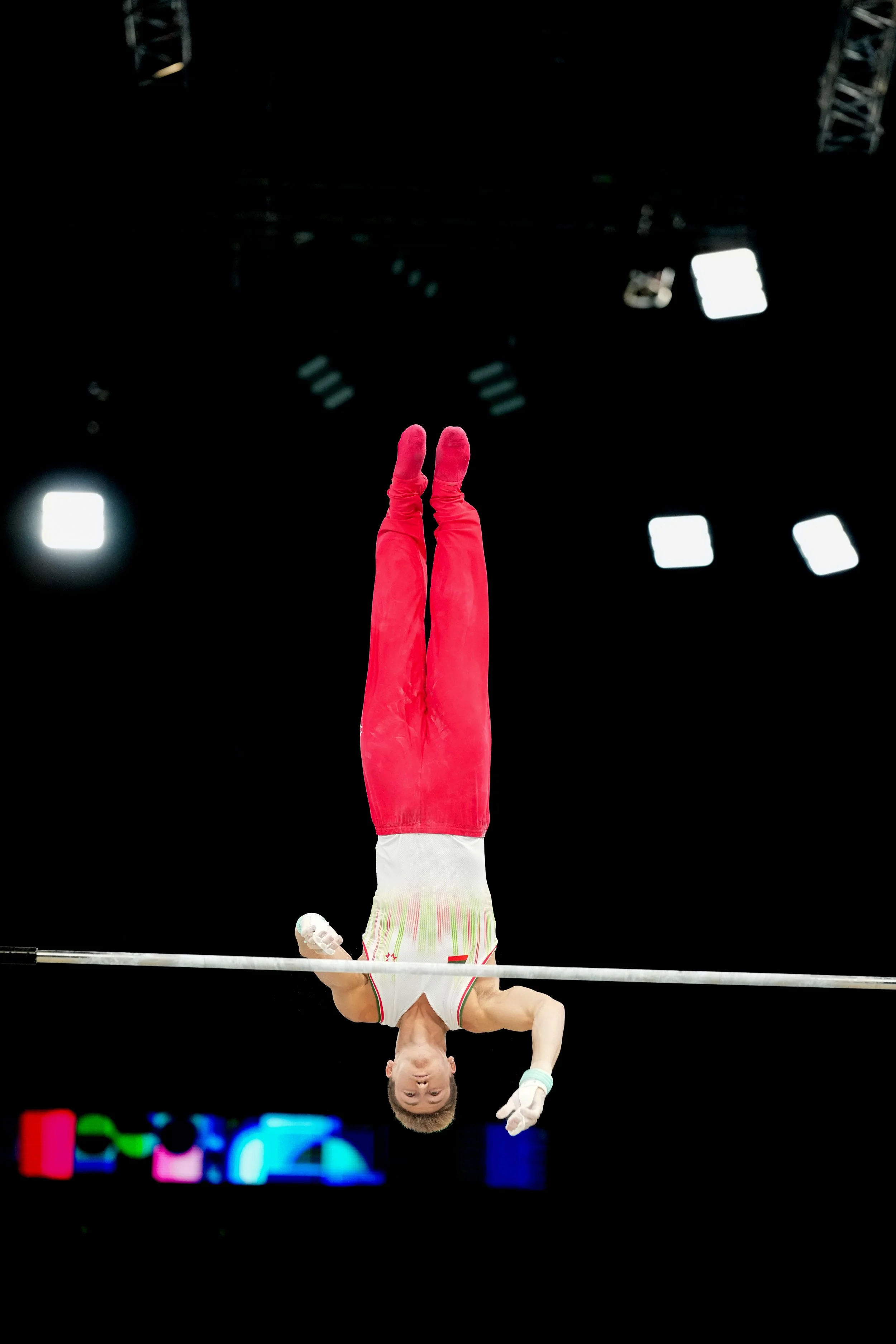Male gymnast performing on the horizontal bar during an international competition in an arena with bright lights and digital screens in the background.