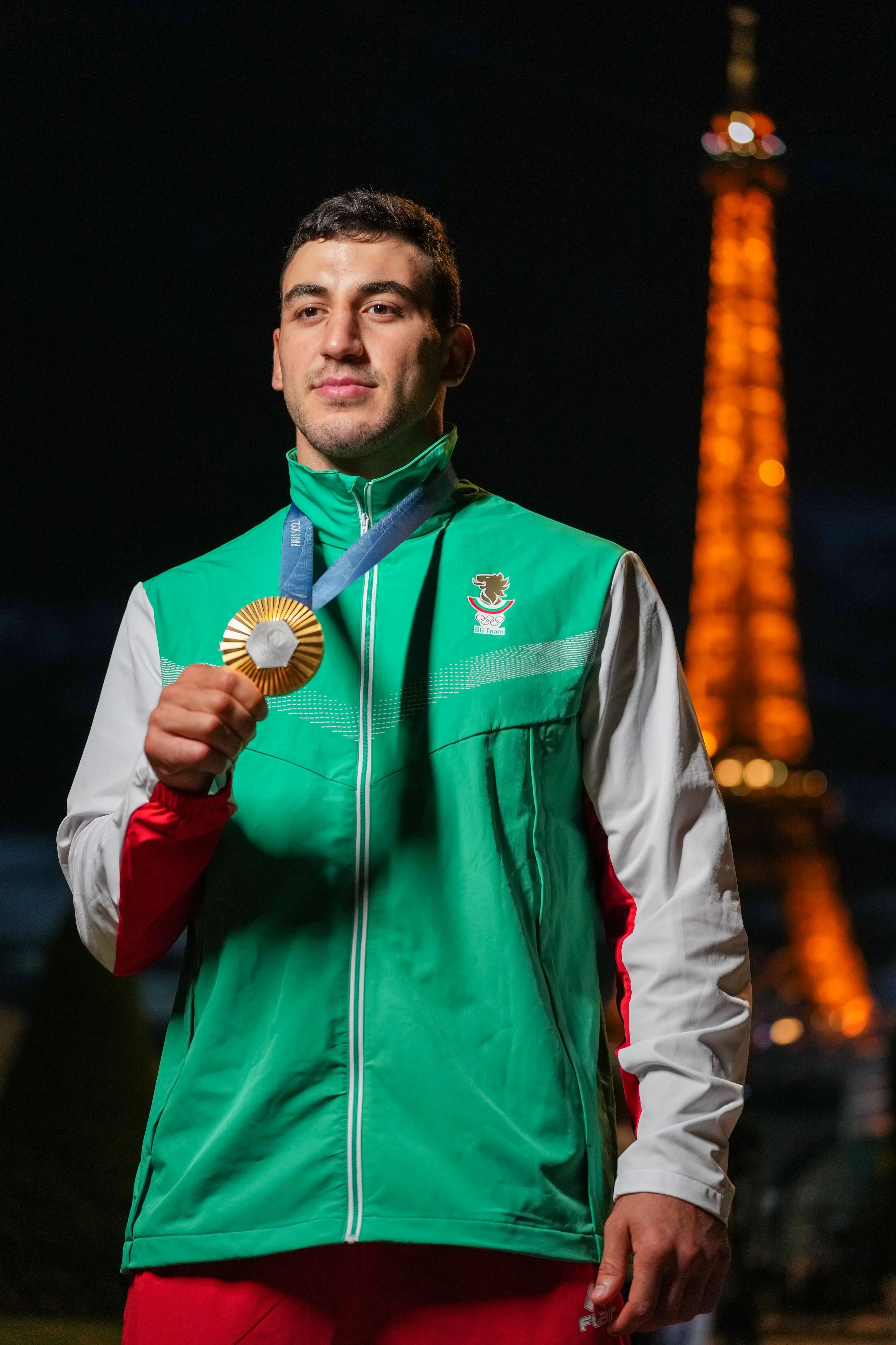 A male athlete stands outdoors at night, holding a gold medal on a blue ribbon, with the Eiffel Tower illuminated in the background.