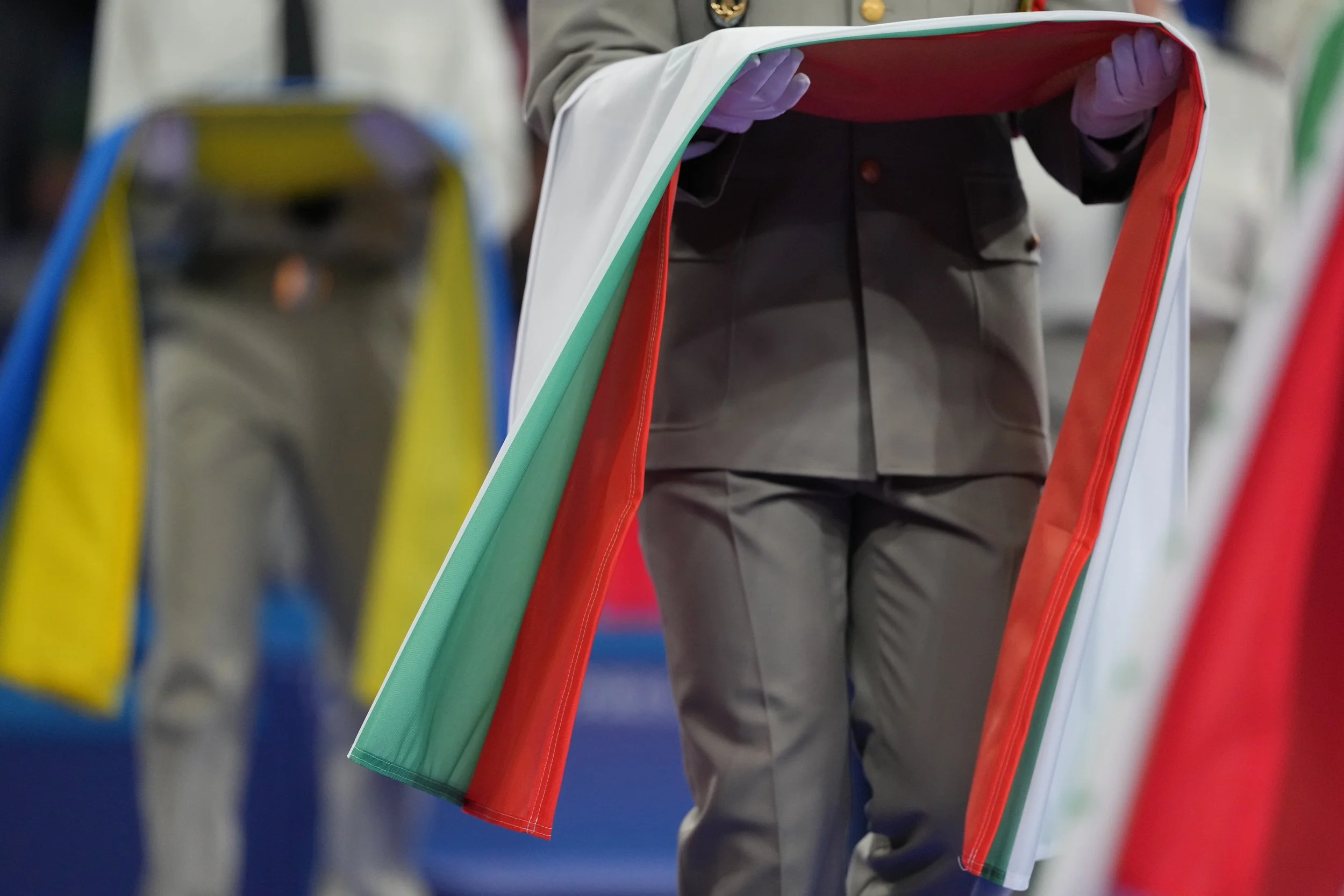 A person wearing a gray suit is holding a large mirror reflecting other suits with flags attached, including the Italian, or Mexican, flags. The background shows colorful flags and blurred figures.