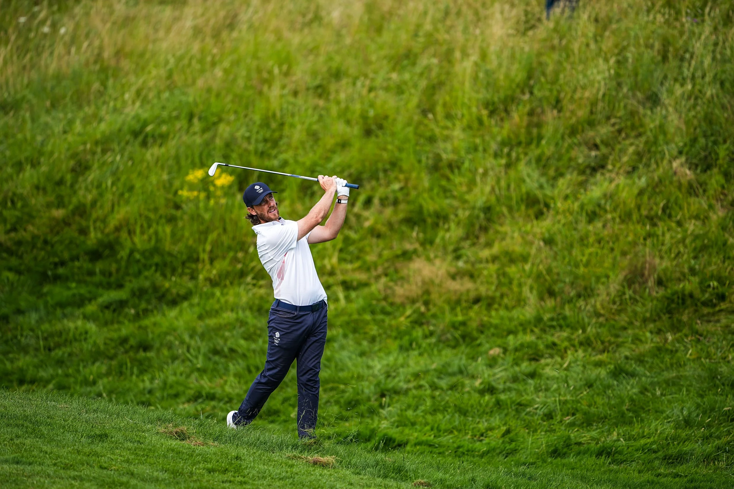A man in a white shirt and navy pants, wearing a navy cap, swings a golf club on a grassy golf course.