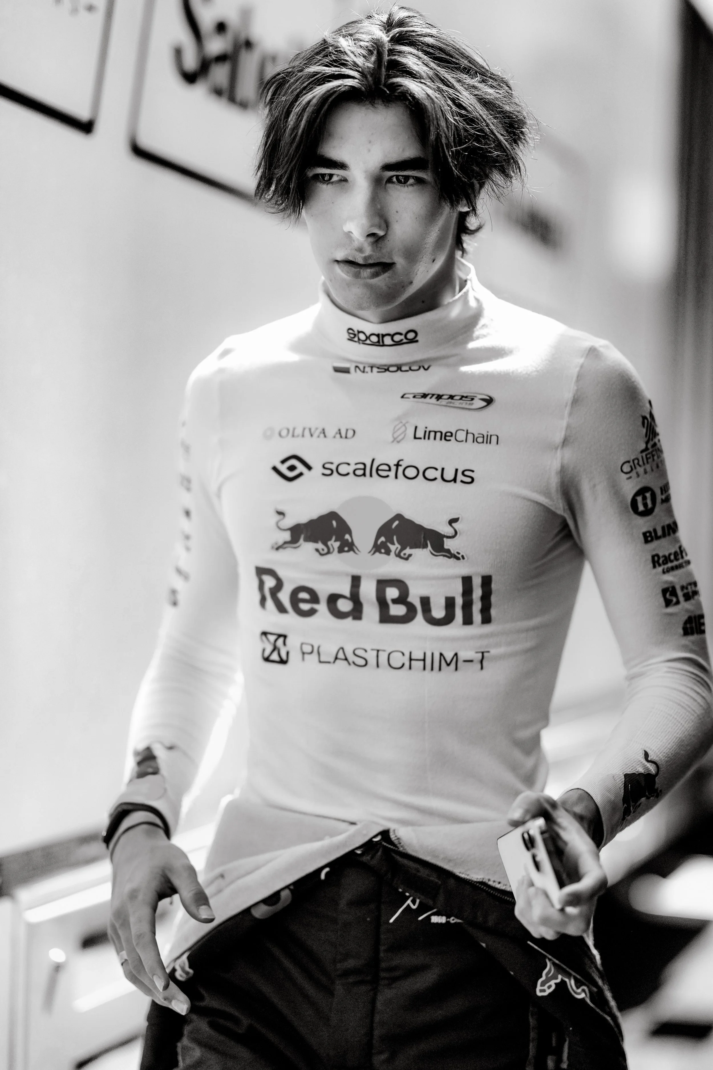 A male race car driver in racing gear, holding a phone, standing in a corridor.