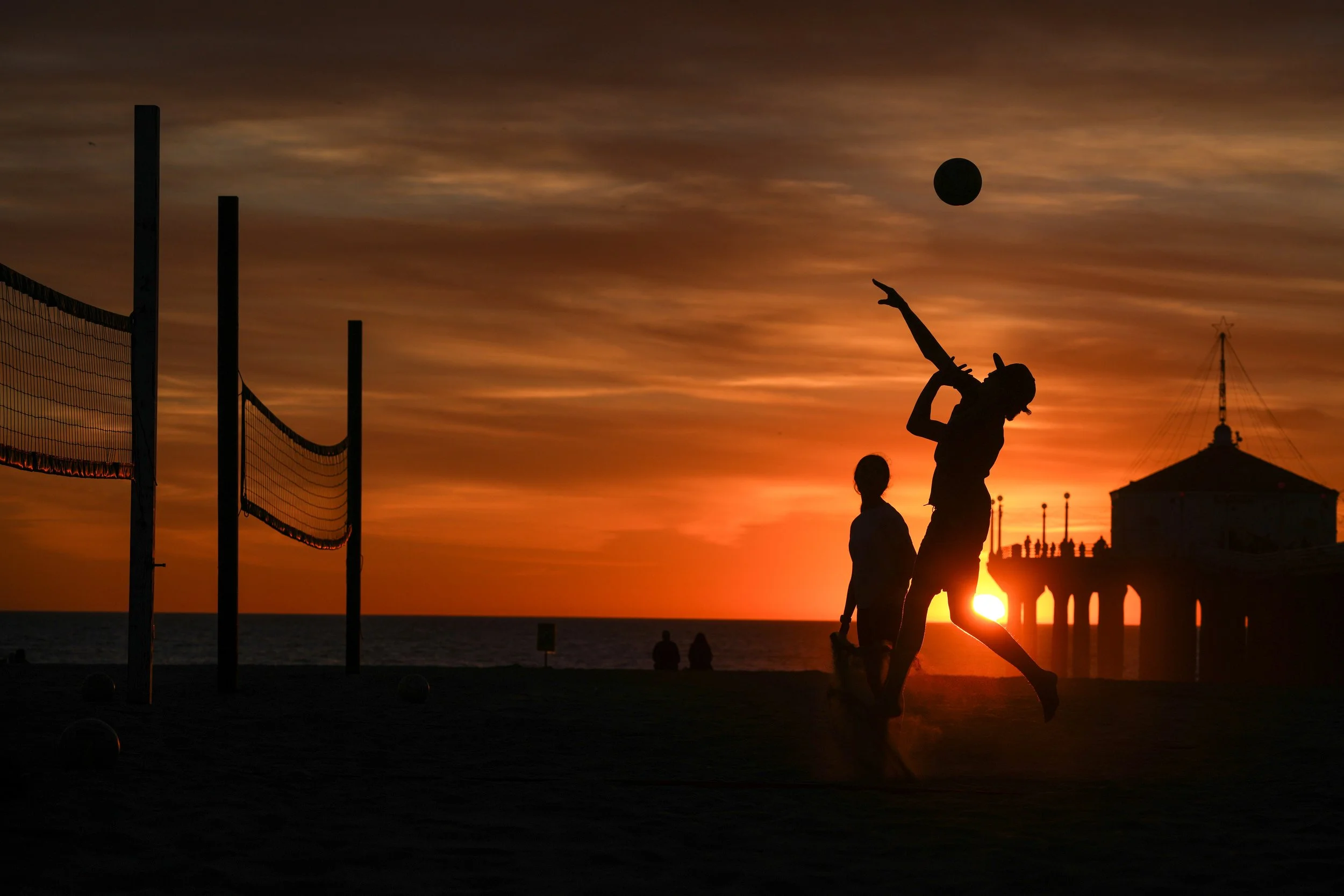 Silhouettes of people on a beach at sunset, one holding a paddle and jumping, with volleyball nets on the left and a pier with a building on the right in the background.