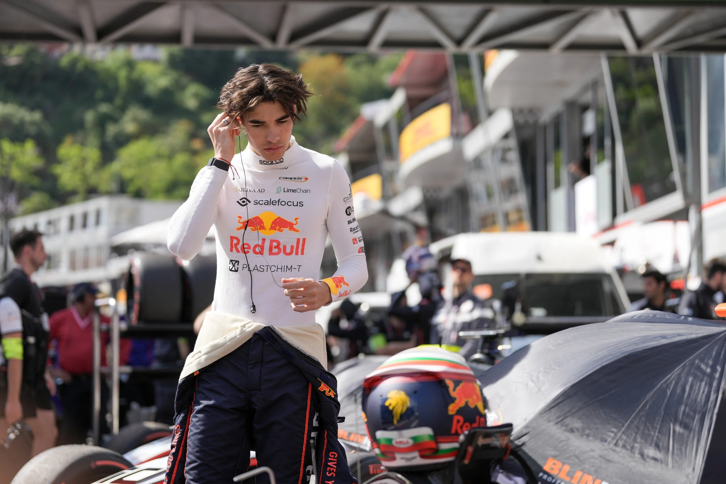 A young male IndyCar driver with messy brown hair, wearing a white racing suit with Red Bull logos, stands next to his race car, adjusting his earpiece and listening to headphones. The background features a racing paddock with crew members, tents, an