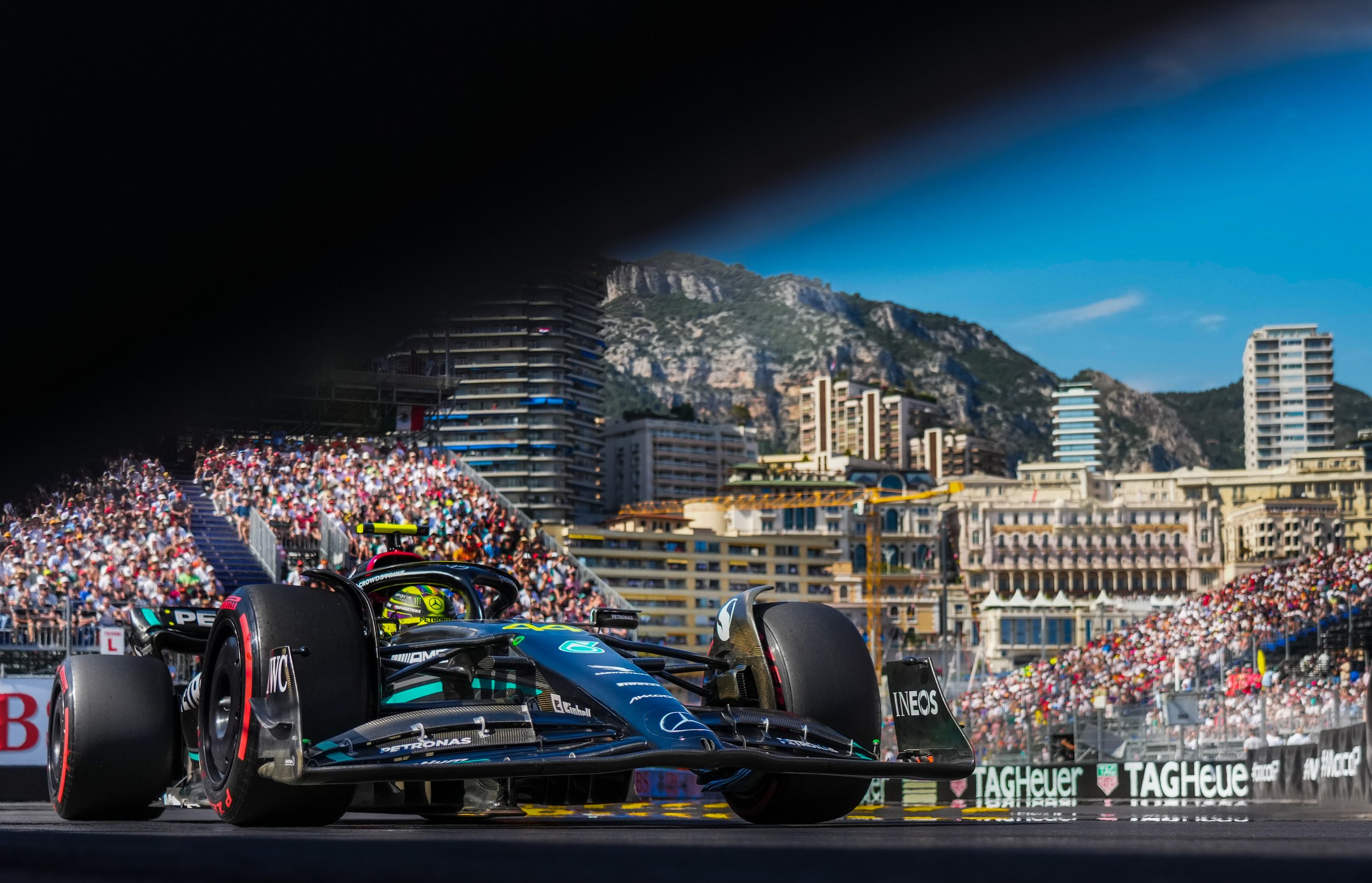 A Formula 1 race car on the track during a race, with a crowd in the grandstands and city buildings and mountains in the background.