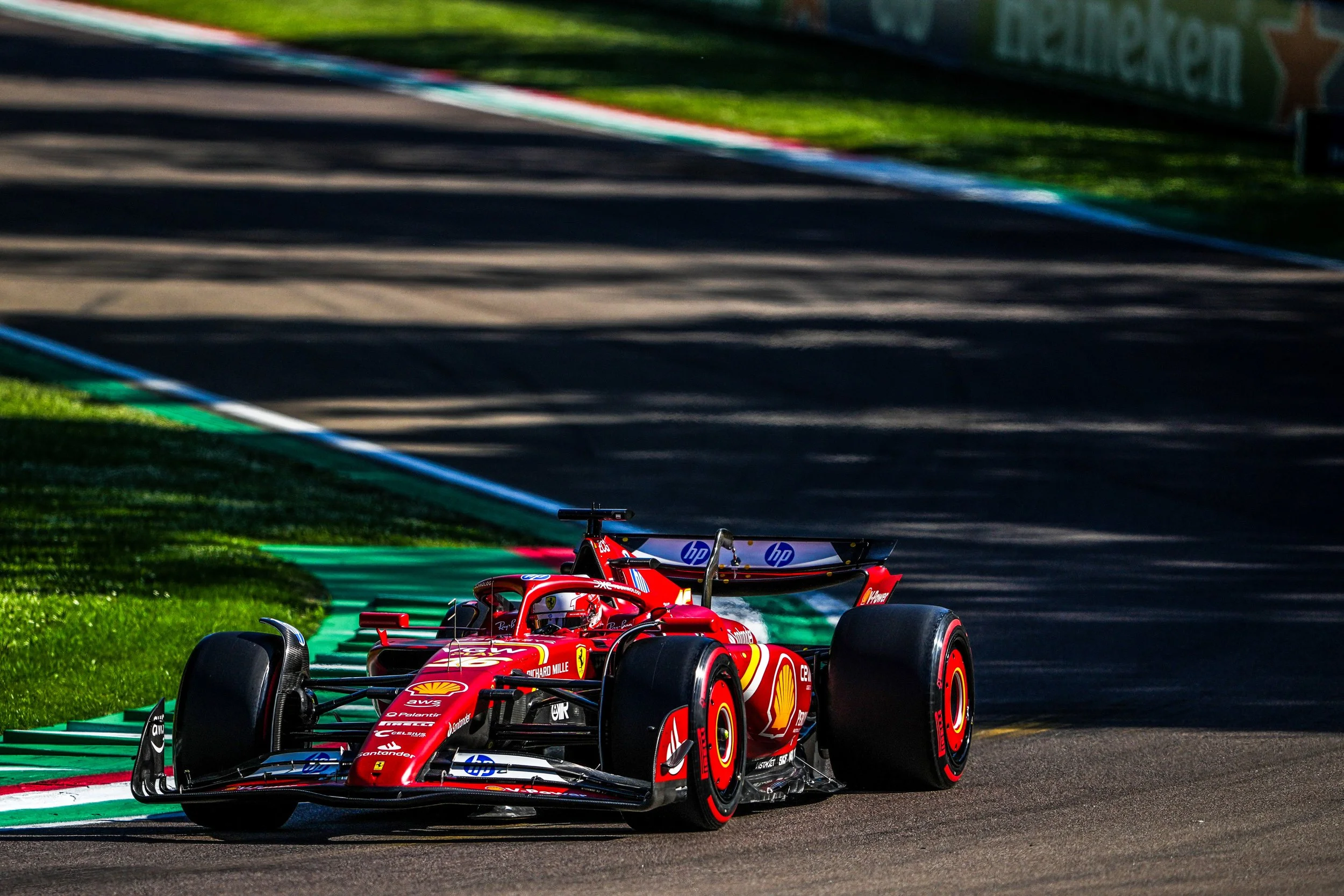 A red Ferrari Formula 1 race car on a race track during daytime, with shadows cast on the asphalt, green grass on the side, and blurred advertisements in the background.