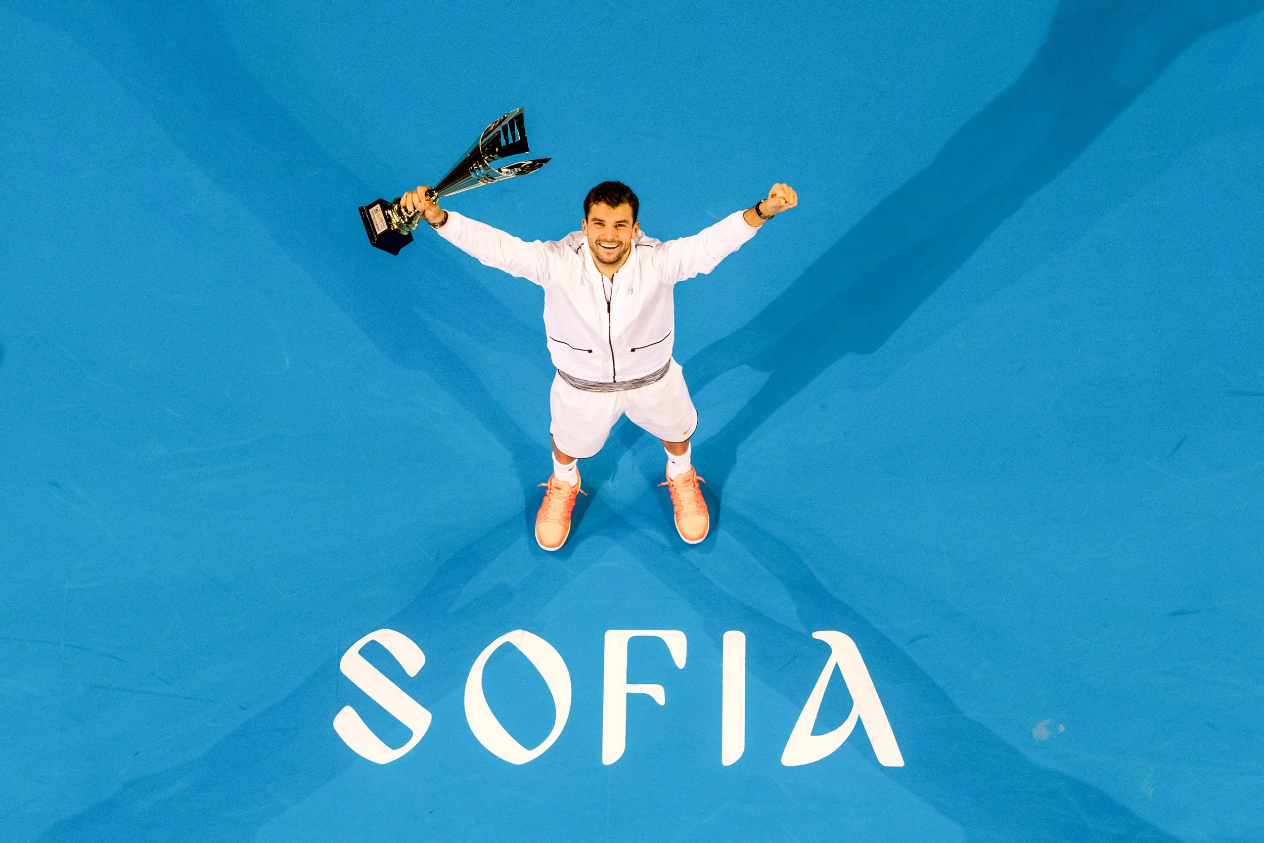 A man standing on a tennis court holding a trophy in his right hand, smiling, with arms outstretched. The court has the brand name 'SOFIA' written on it.