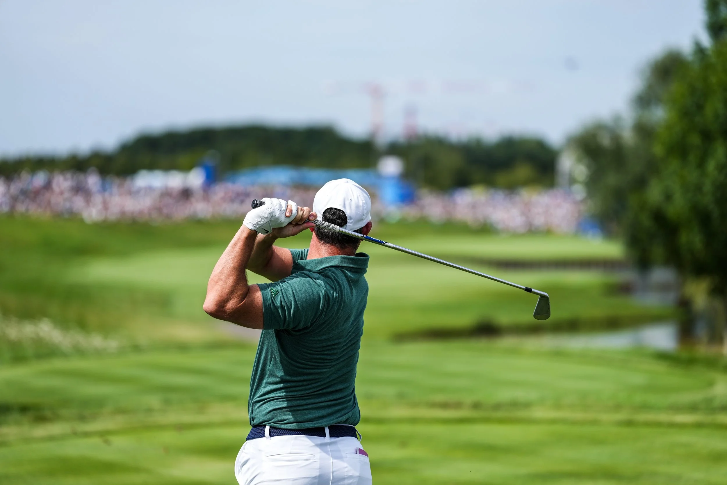 Male golfer in green shirt and white cap swinging a golf club on a lush golf course with a crowd in the background.