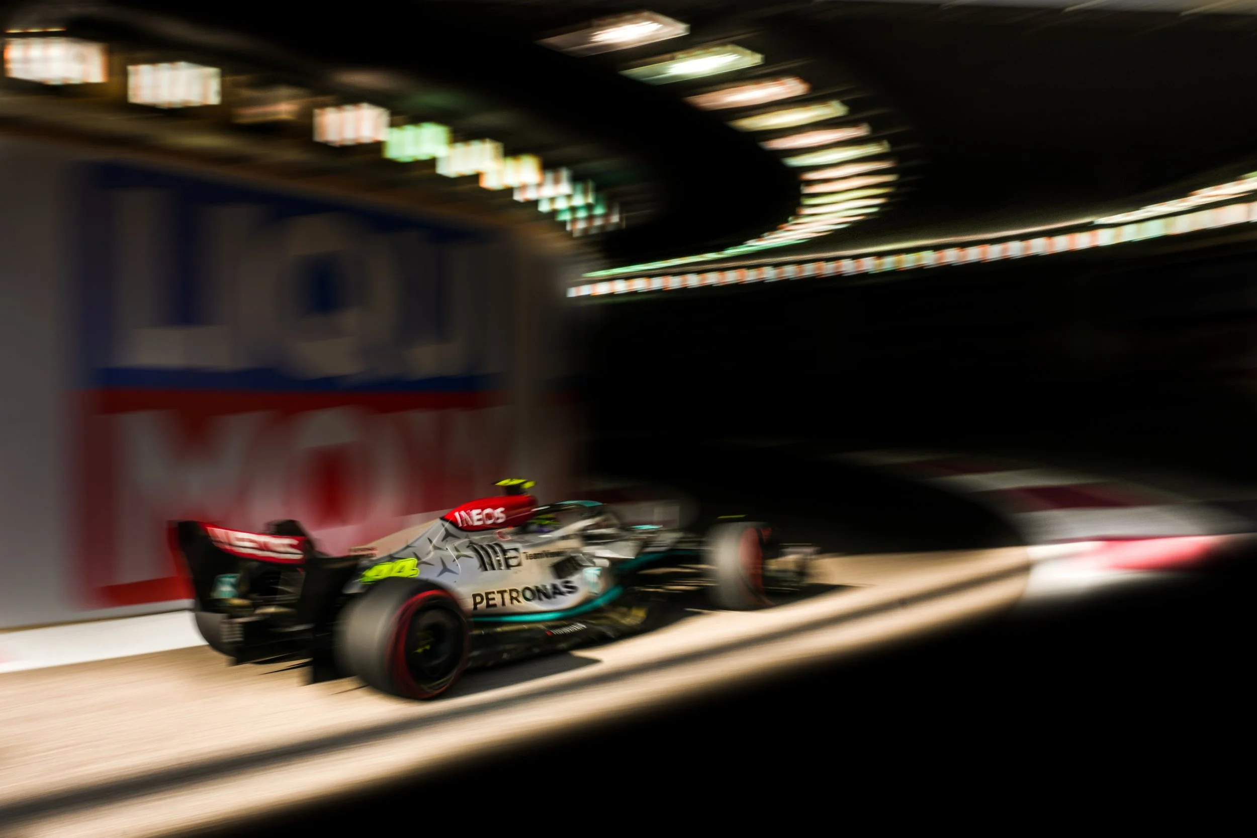 Blurred Formula 1 car racing at night on a track with bright overhead lights.