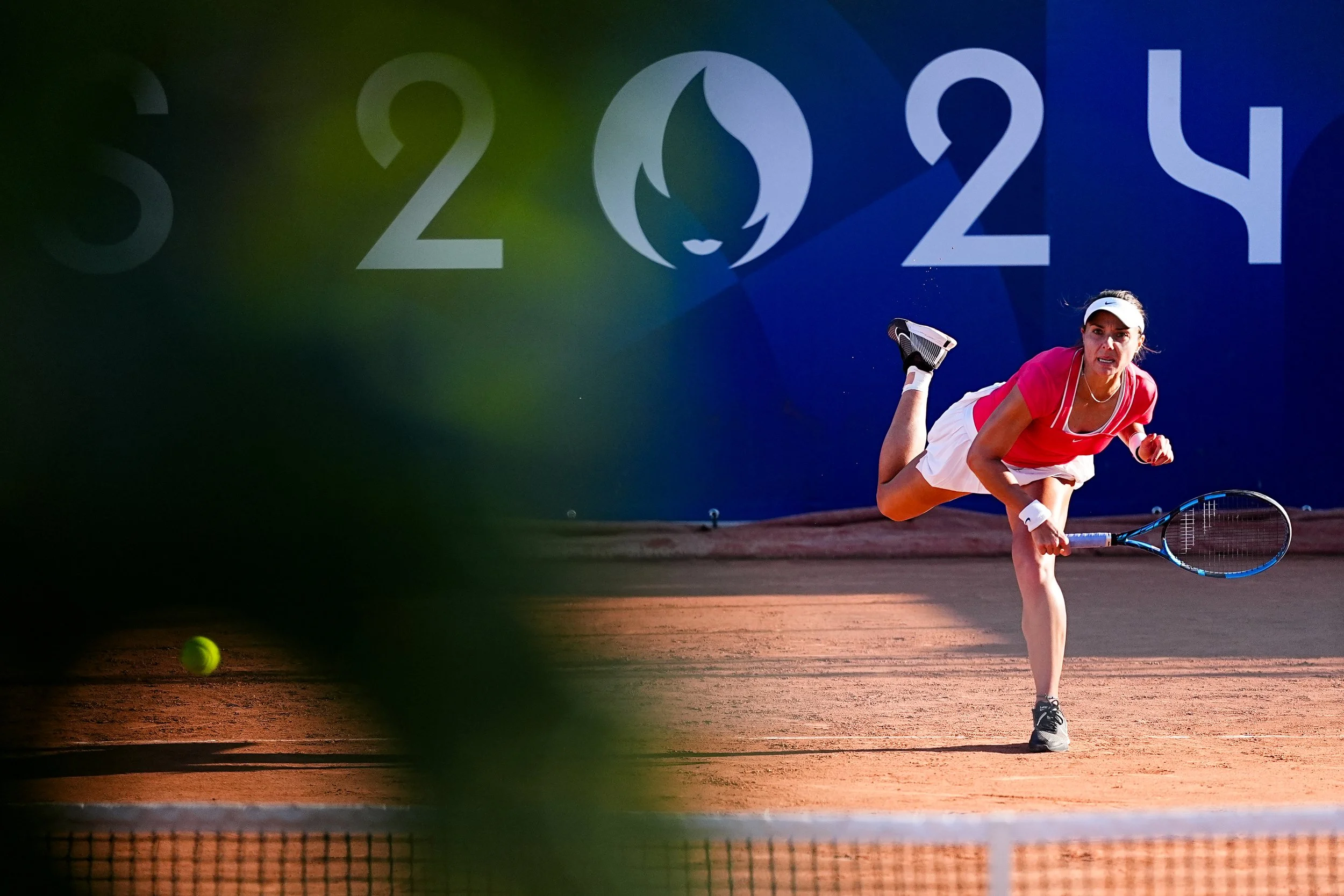 Female tennis player in a pink shirt and white skirt lunges to hit a ball on a clay court during a match, with a large blue background displaying the numbers 2024 and a flame logo.