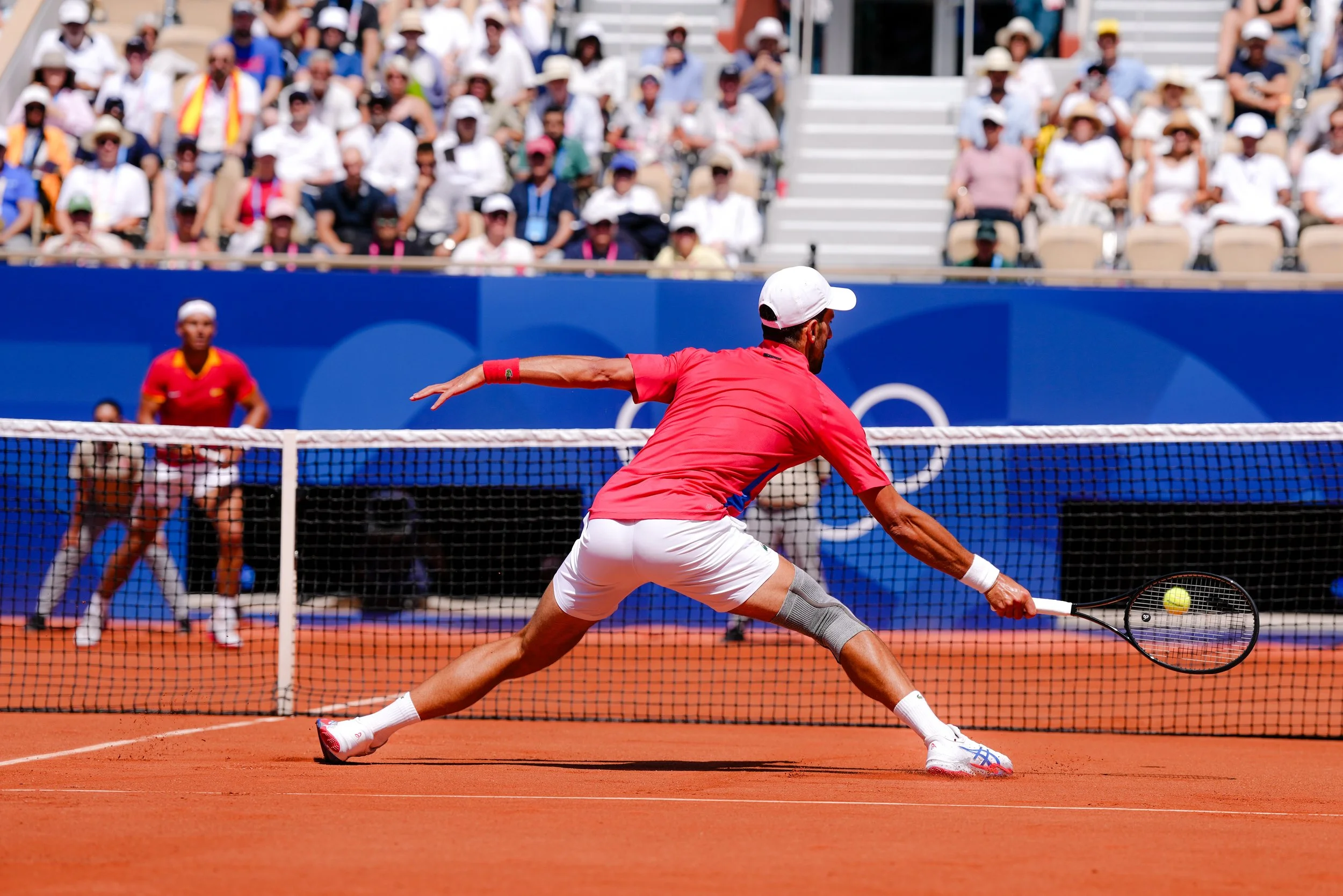 A tennis player in a red shirt and white shorts reaching to hit a tennis ball with a racket on a clay court, with a crowd sitting in the stands behind him.