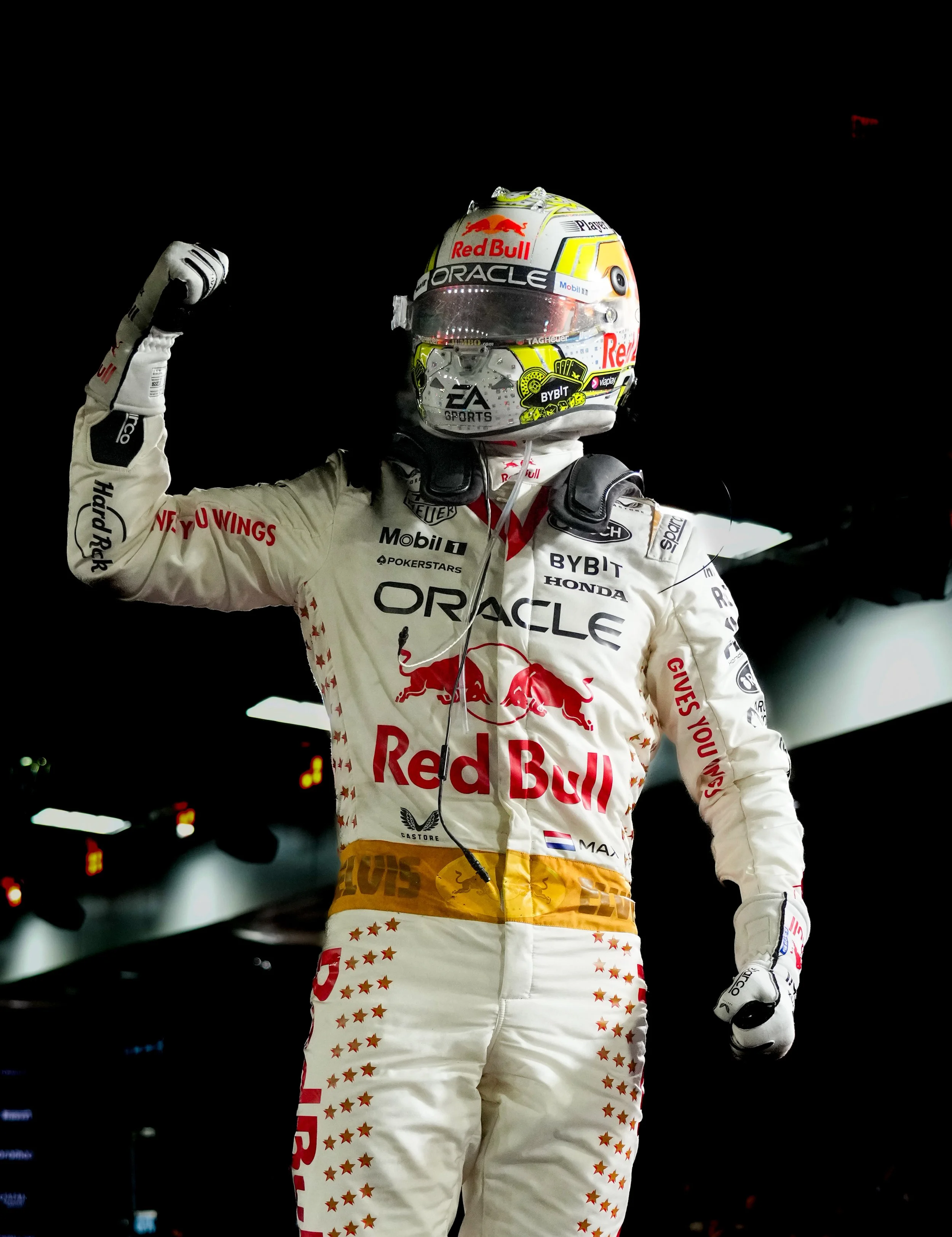 Race car driver in racing suit and helmet celebrating with a flexed arm in a dimly lit garage or pit area.