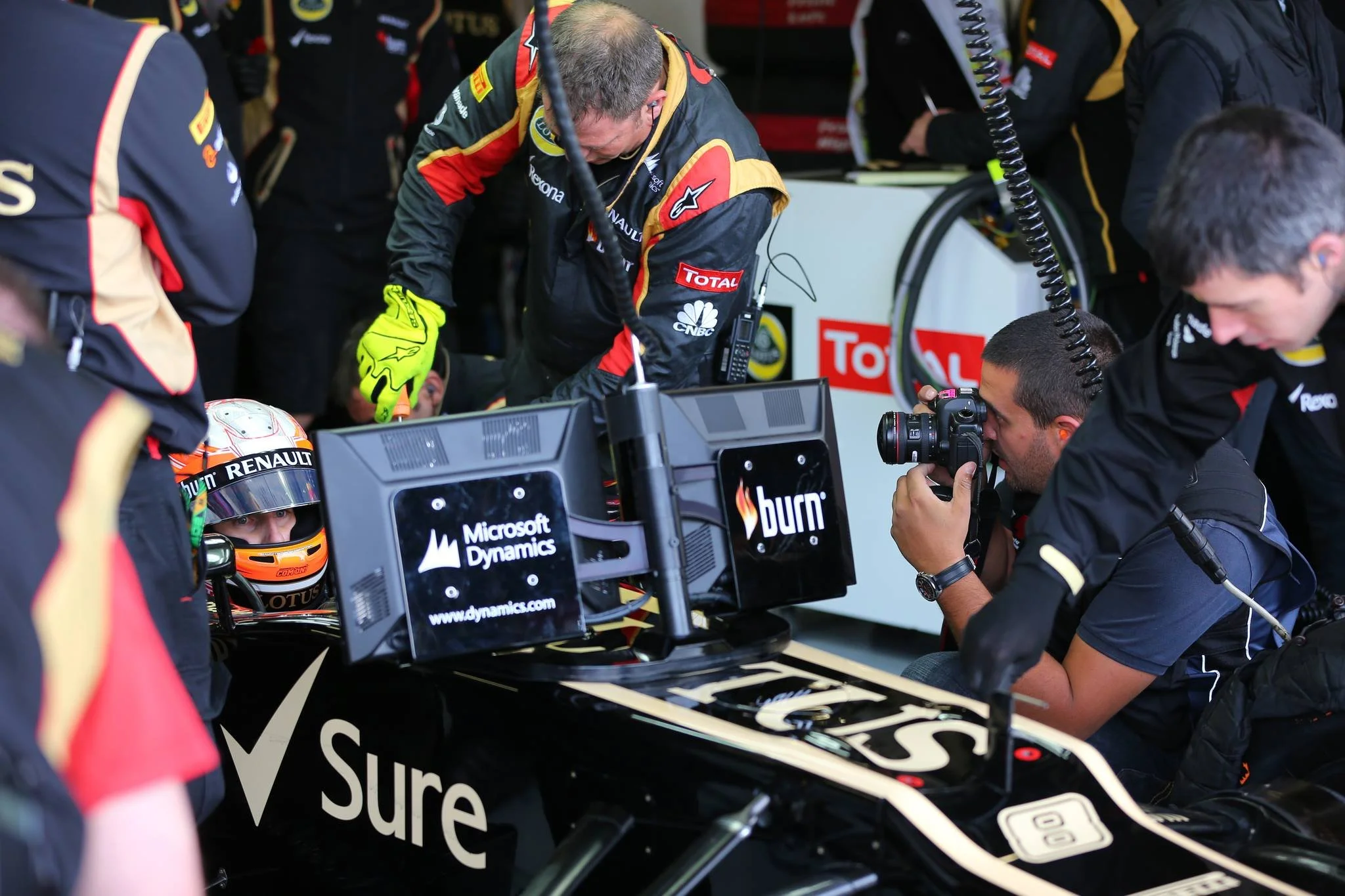 Formula 1 race car with drivers and team members preparing in a garage, with a photographer taking pictures.