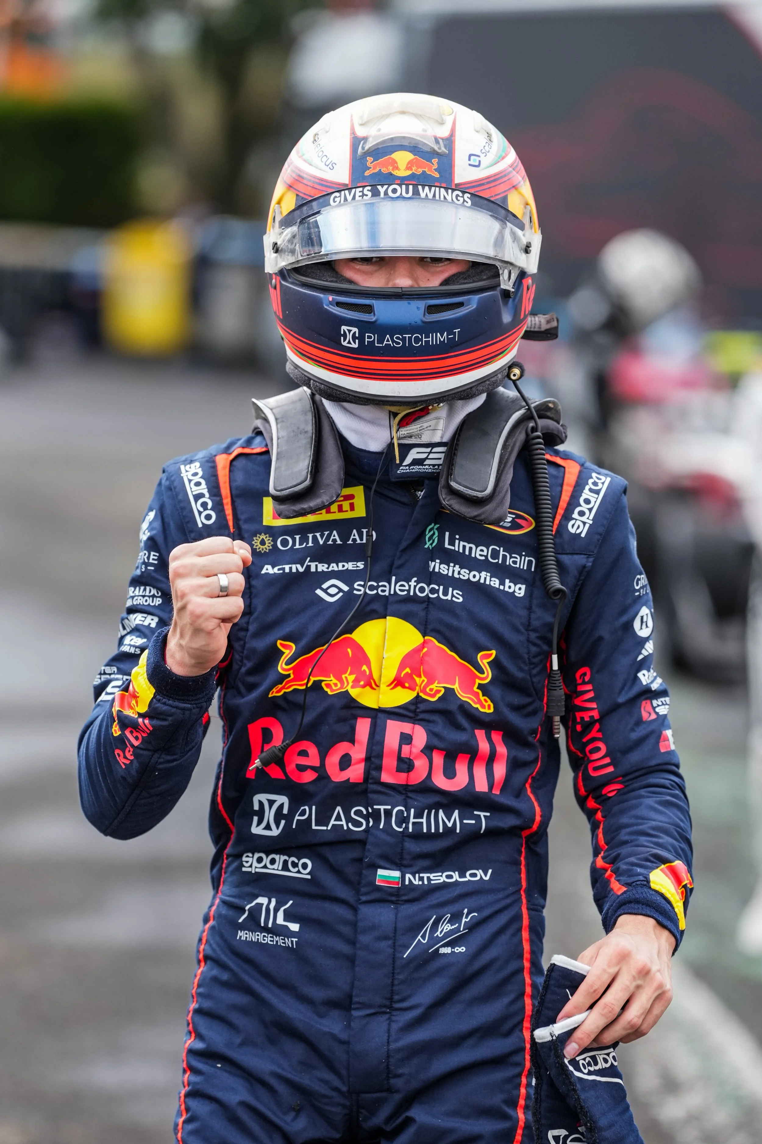 A race car driver in a blue racing suit with Red Bull sponsorship, wearing a full-face helmet with a visor, standing outdoors, giving a fist bump gesture.