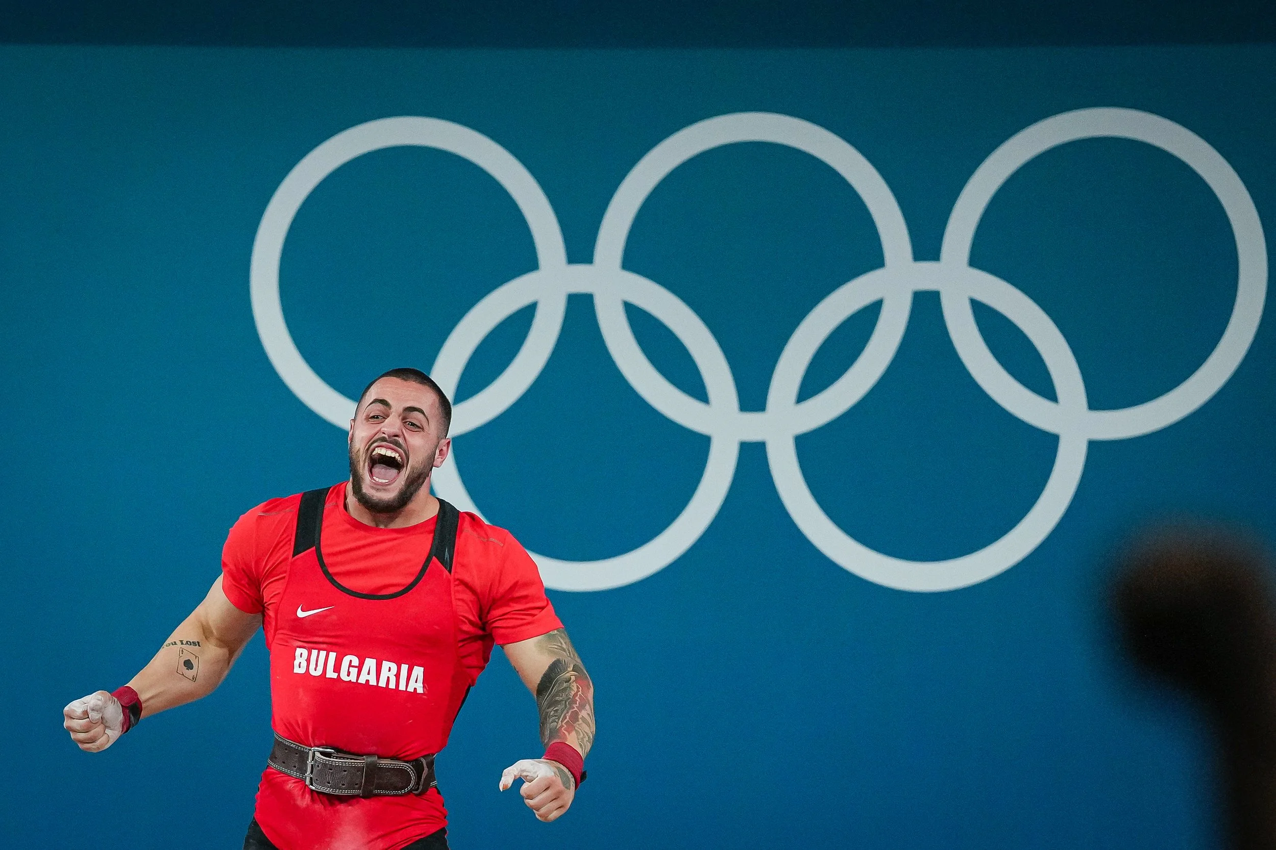 A male athlete in a red Bulgaria shirt celebrating in front of a large Olympic rings logo.