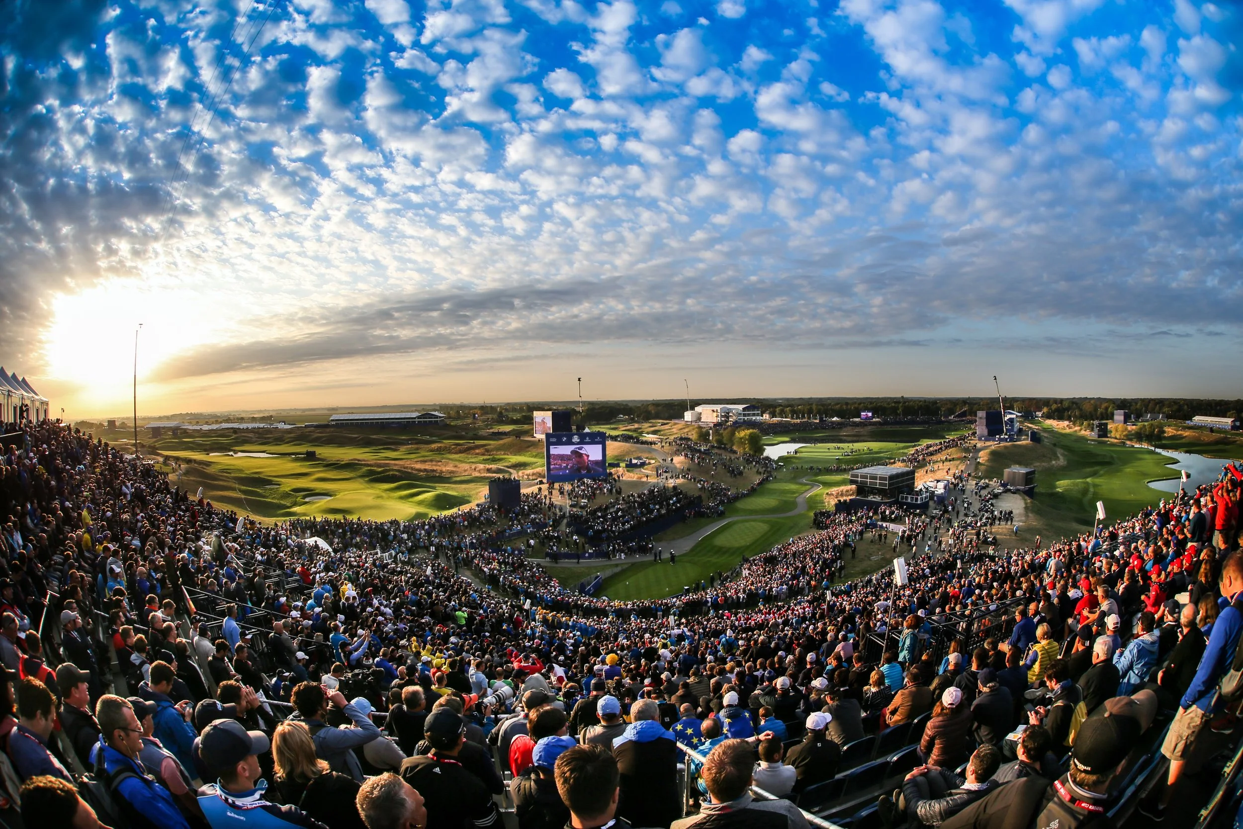 A large crowd of spectators watching a golf tournament at a golf course during sunset.