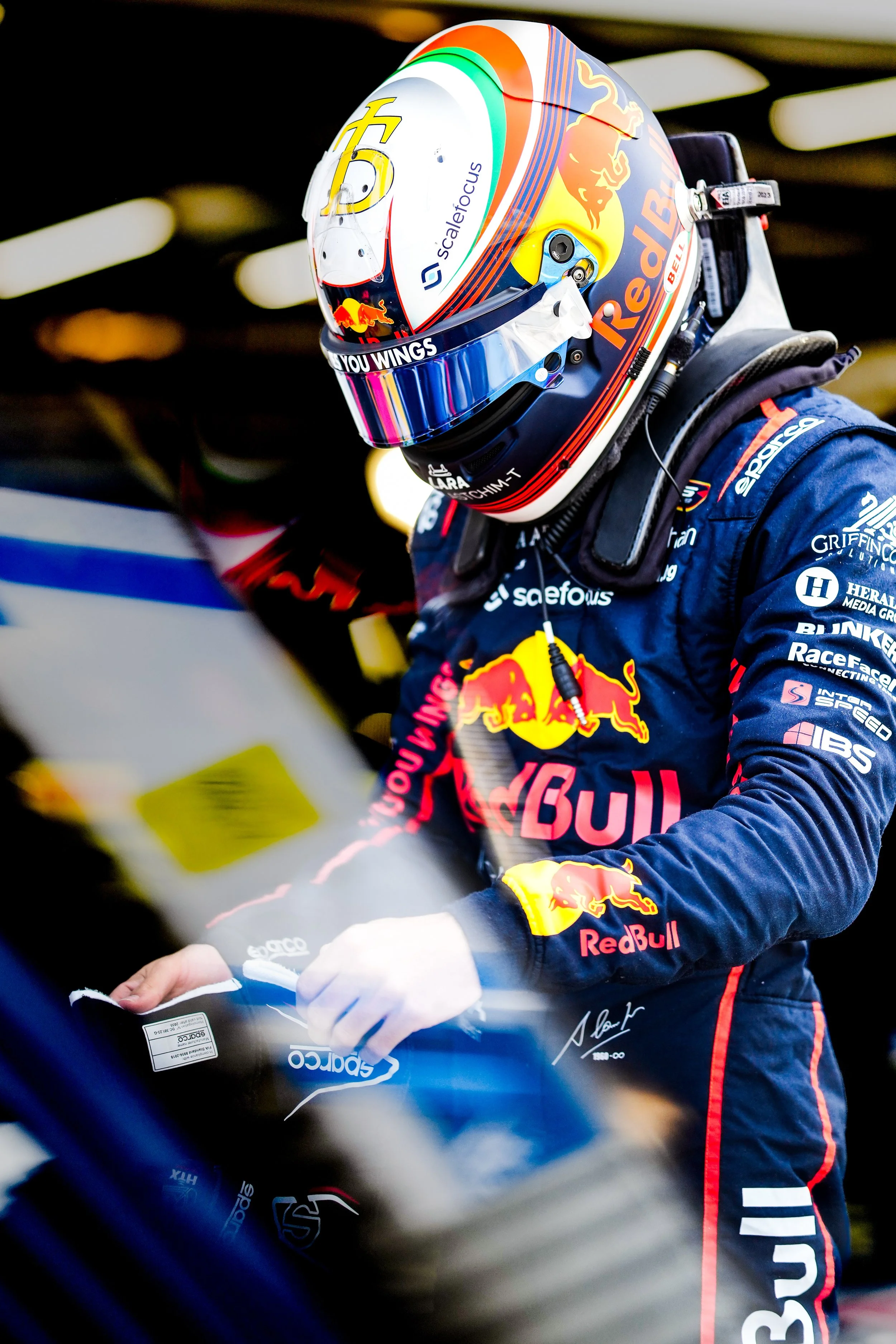 A race car driver in racing gear and a Red Bull helmet stands next to a racing vehicle, likely preparing for a race.