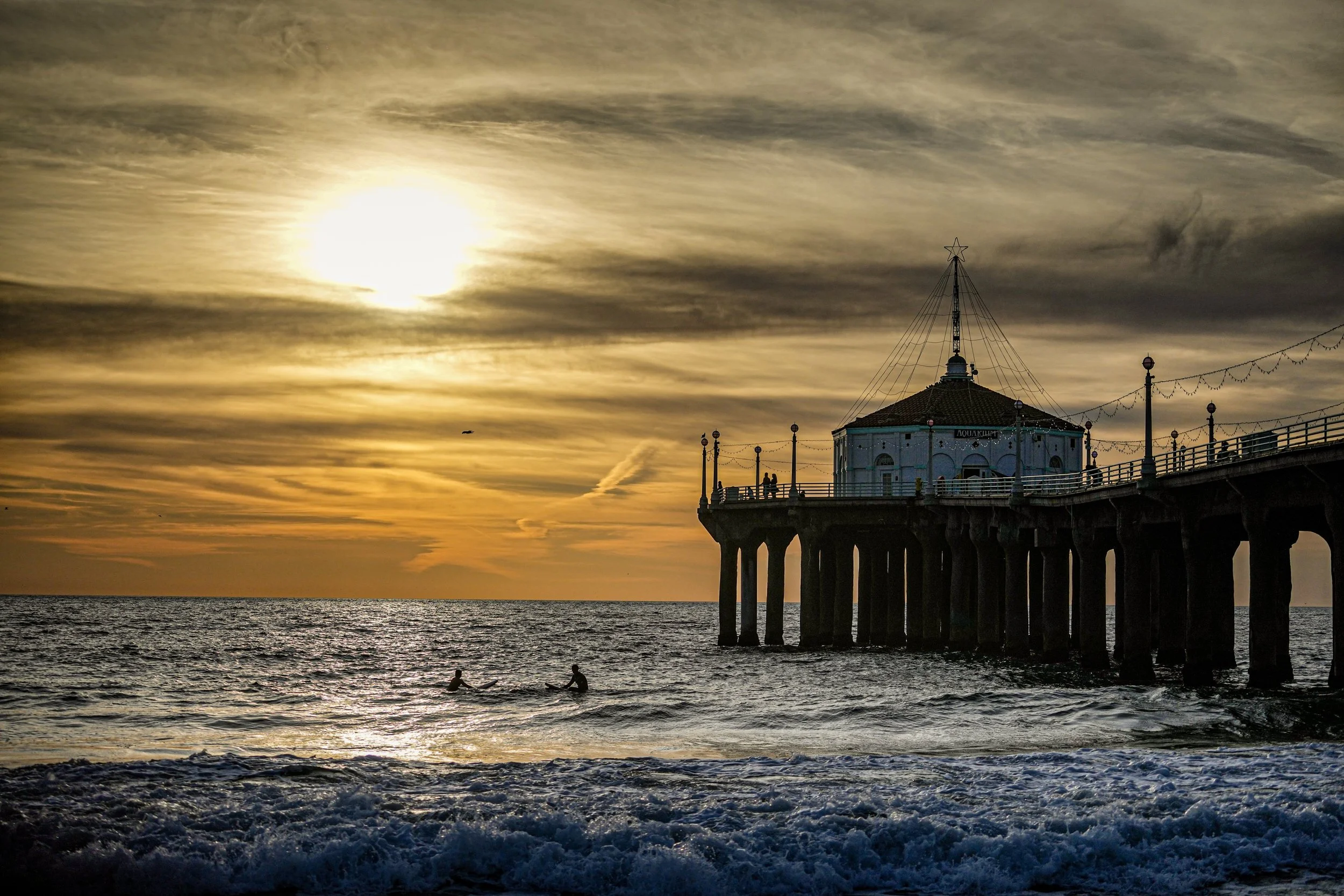 Sunset over the ocean with a pier extending into the water, silhouetted structure on the pier, two people swimming in the water, and cloudy sky overhead.