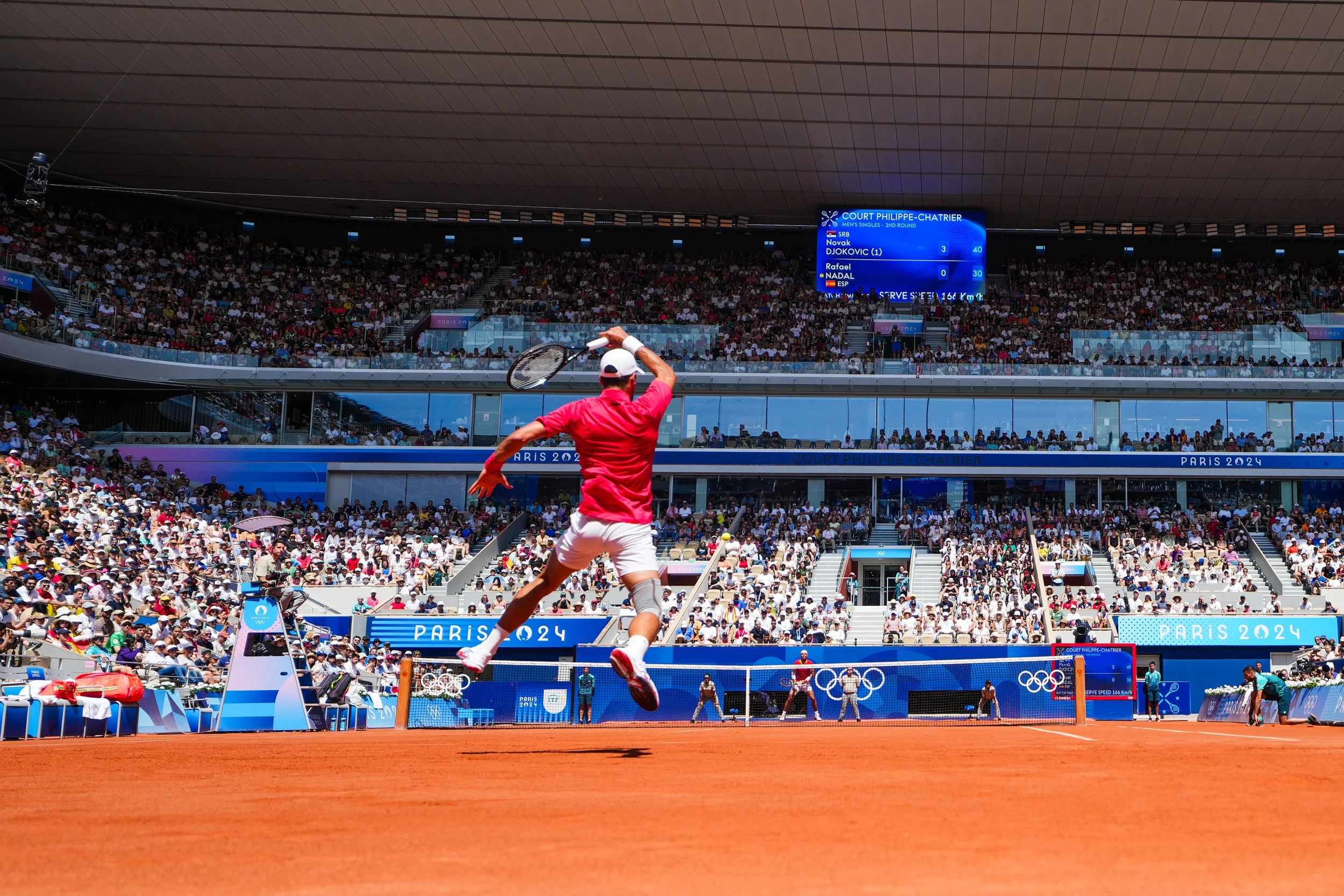 A tennis player in red shirt and white shorts preparing to hit a tennis ball during a match at the Paris 2024 Olympics with a large crowd in the stands and a scoreboard overhead.