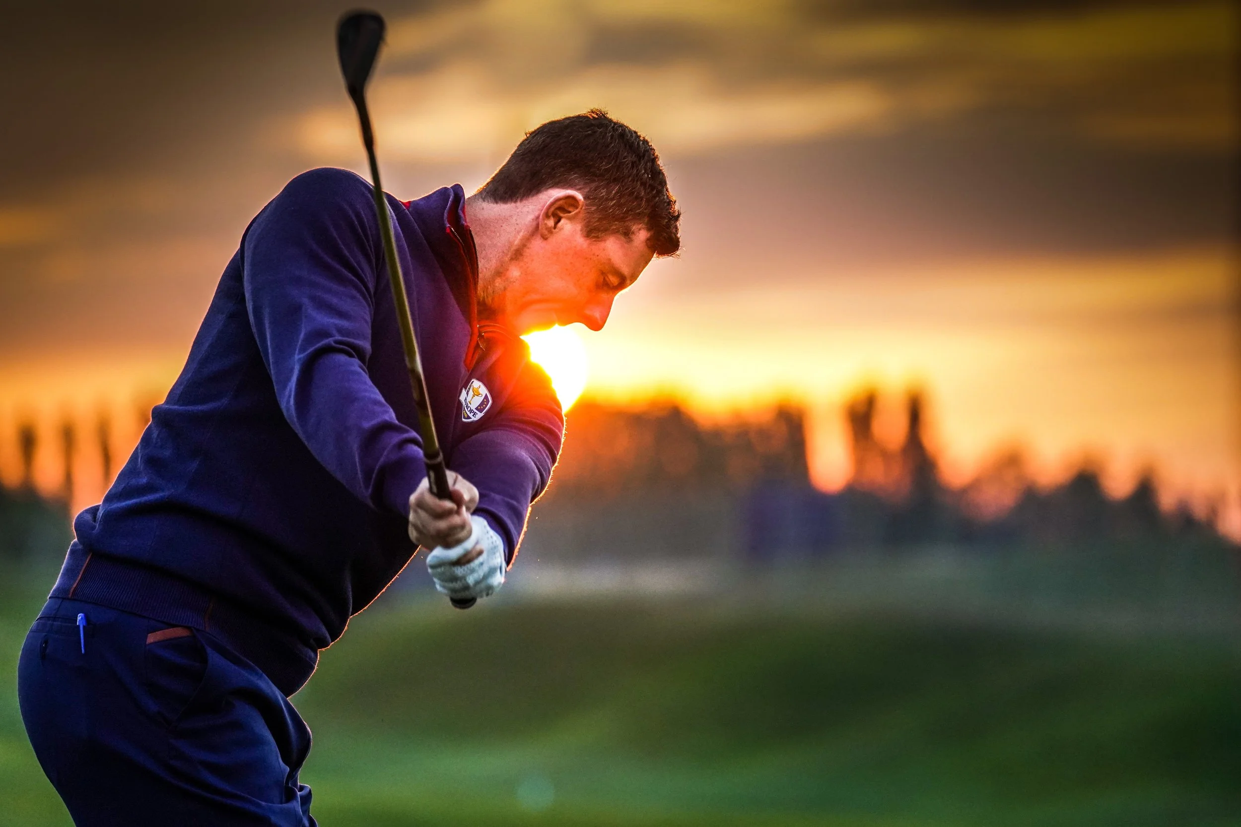 A man playing golf during sunset, swinging a club on the golf course.