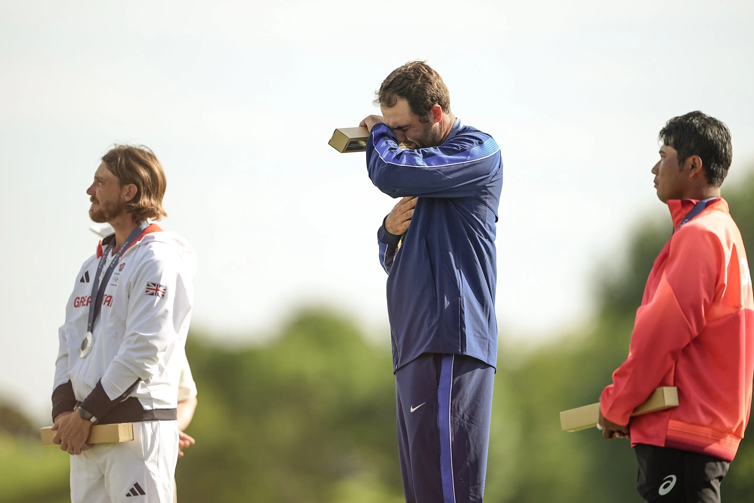 Three male athletes standing outdoors on a podium, holding medals and facing different directions. The athlete in the middle is crying and holding a gold medal, wearing a blue sports outfit.