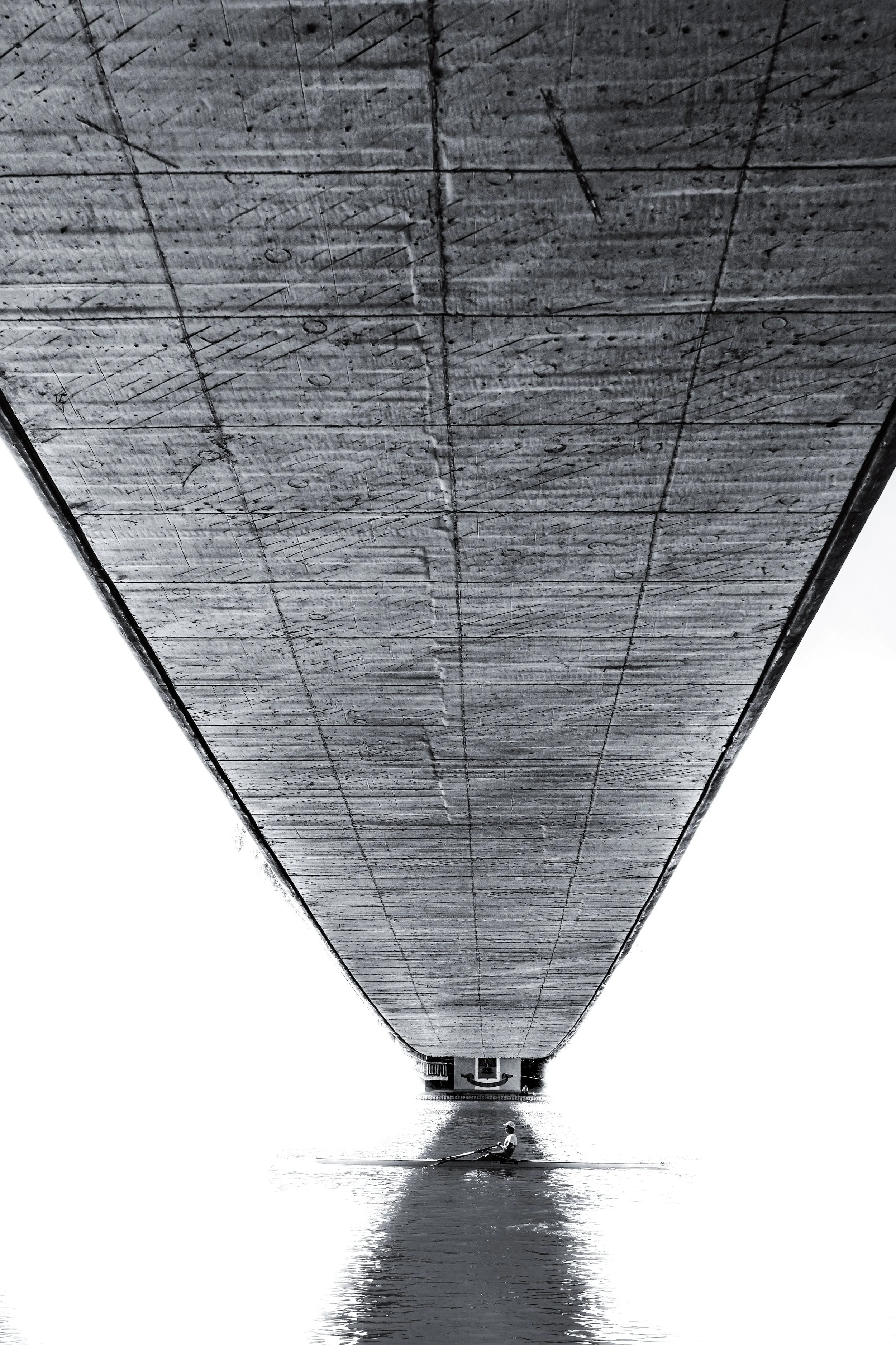 A person rowing a boat under a large concrete bridge above the water.