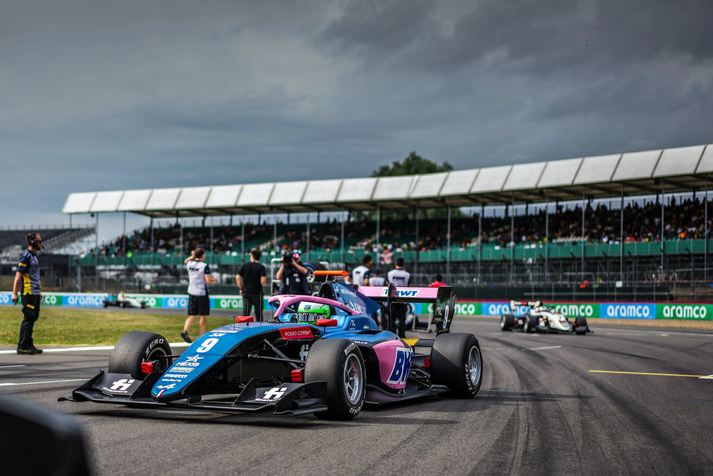 Race cars on a racetrack with team members and spectators in the background under a cloudy sky.