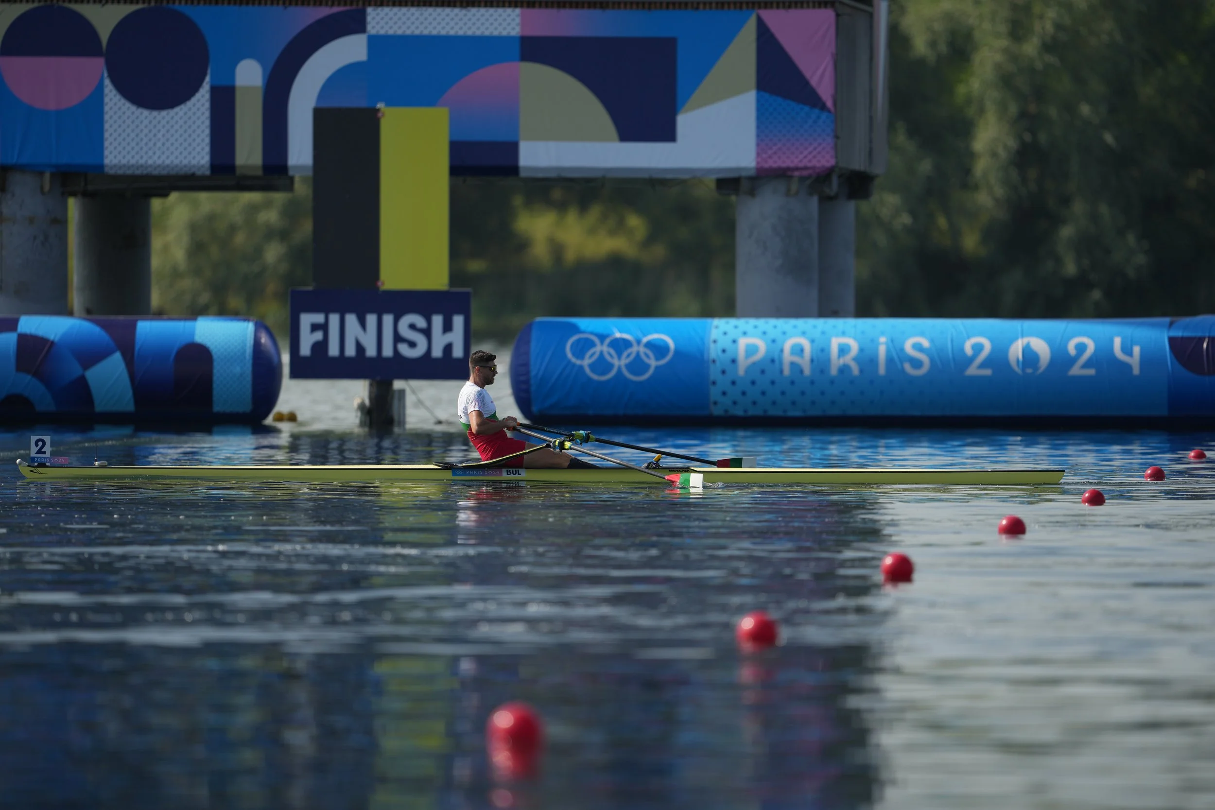 A male athlete in a single rowing boat racing on water with red buoys marking the lane, near a finish line with Olympic rings, indicating a competition during the Paris 2024 Olympics.