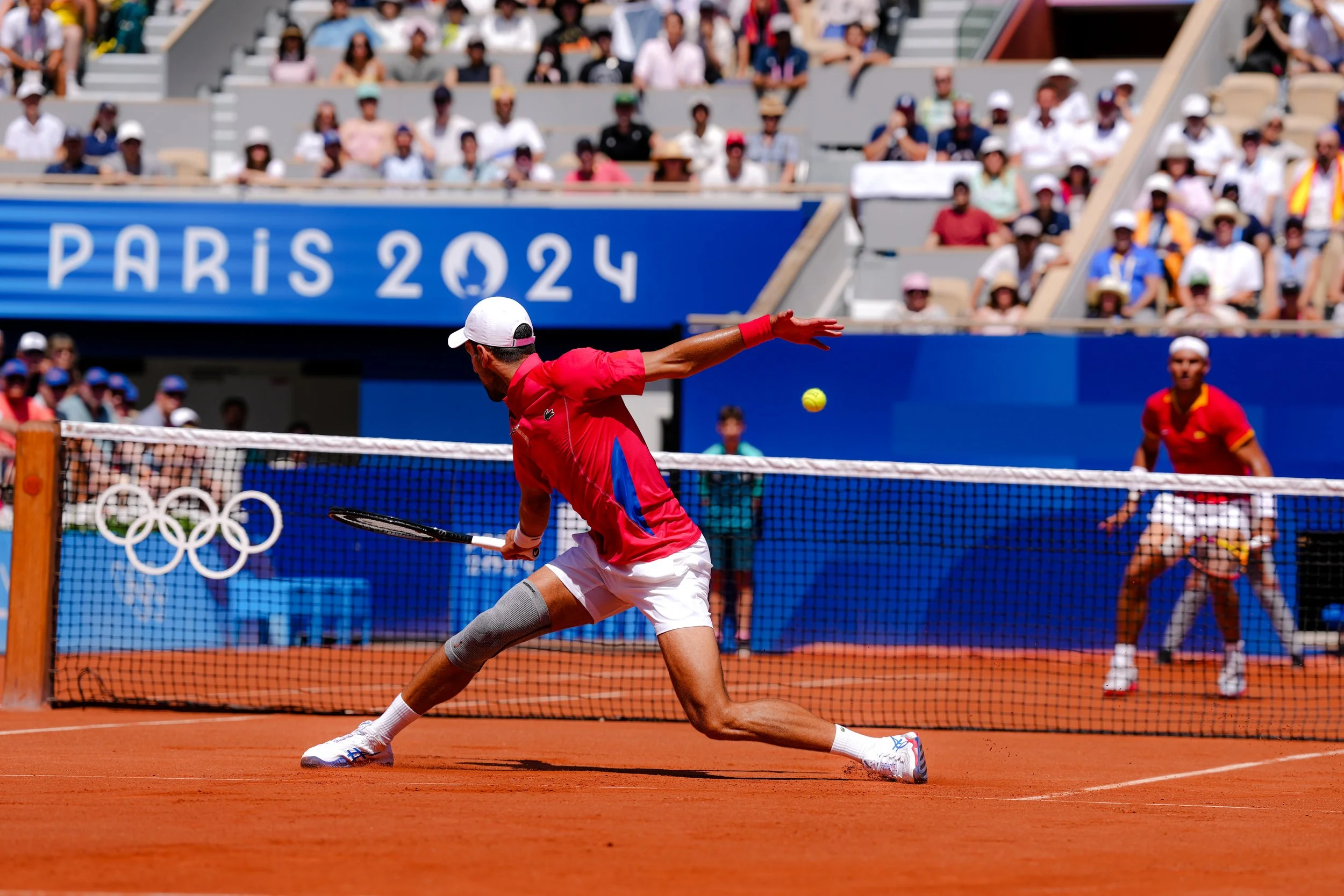 Tennis players in action during a match at the Paris 2024 Olympics, with one player stretching to return the ball and a crowd watching from the stands.