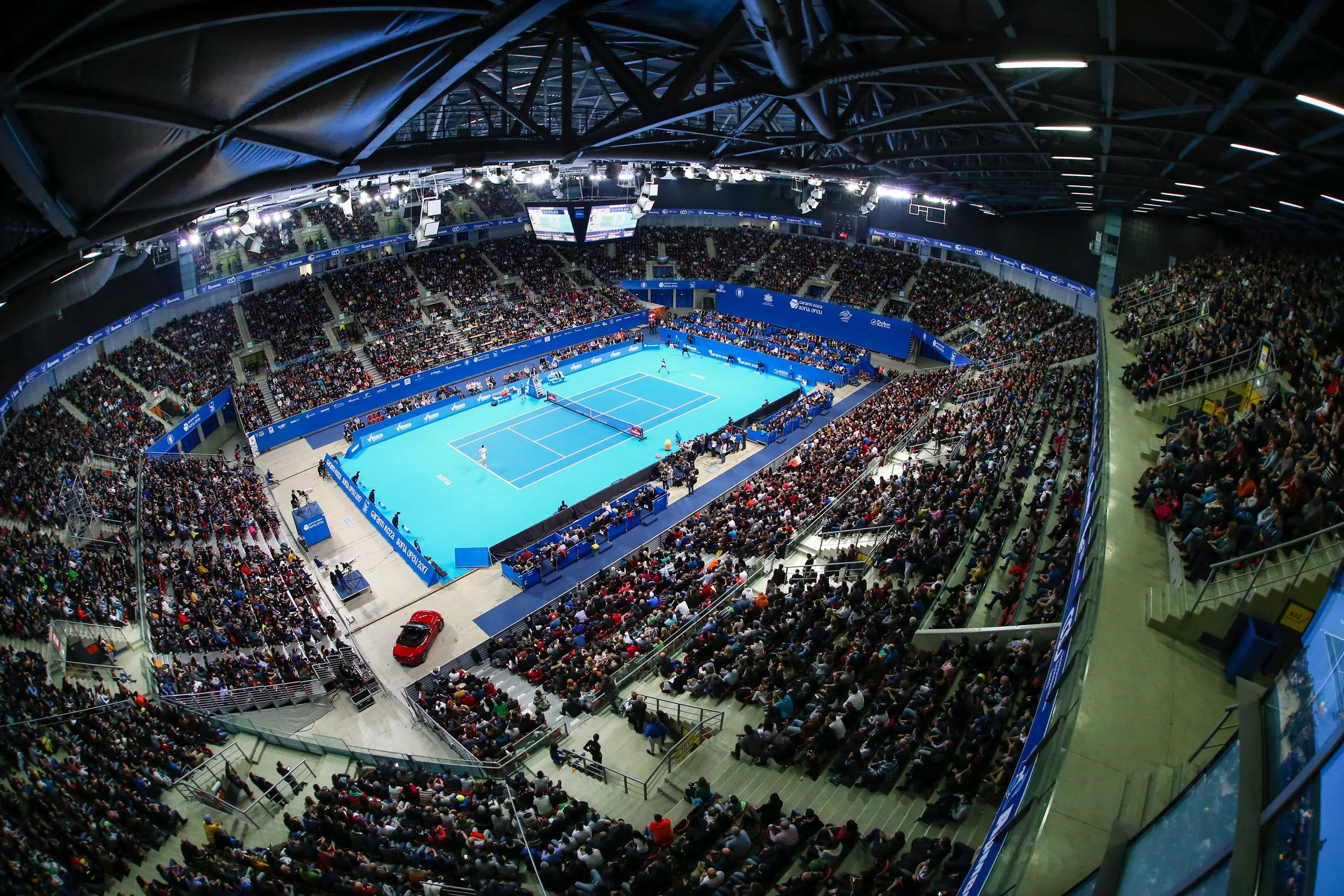 An indoor tennis arena filled with spectators watching a match on a blue court, with large screens displaying the game.