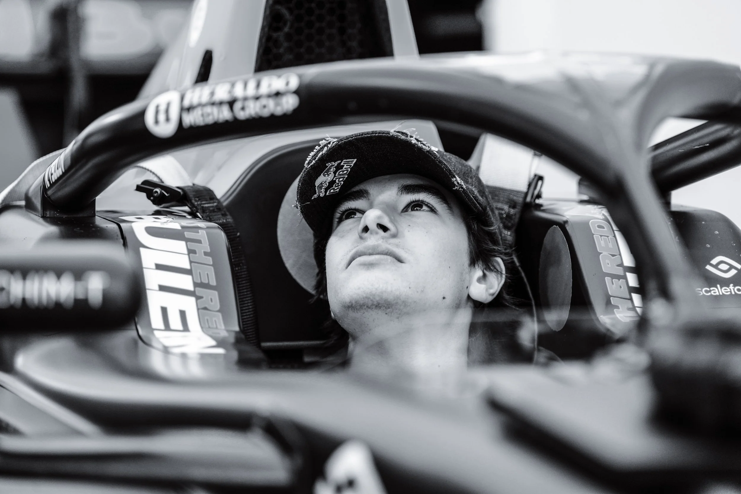 Young man in racing car cockpit, looking upward, wearing a cap, black and white photo.
