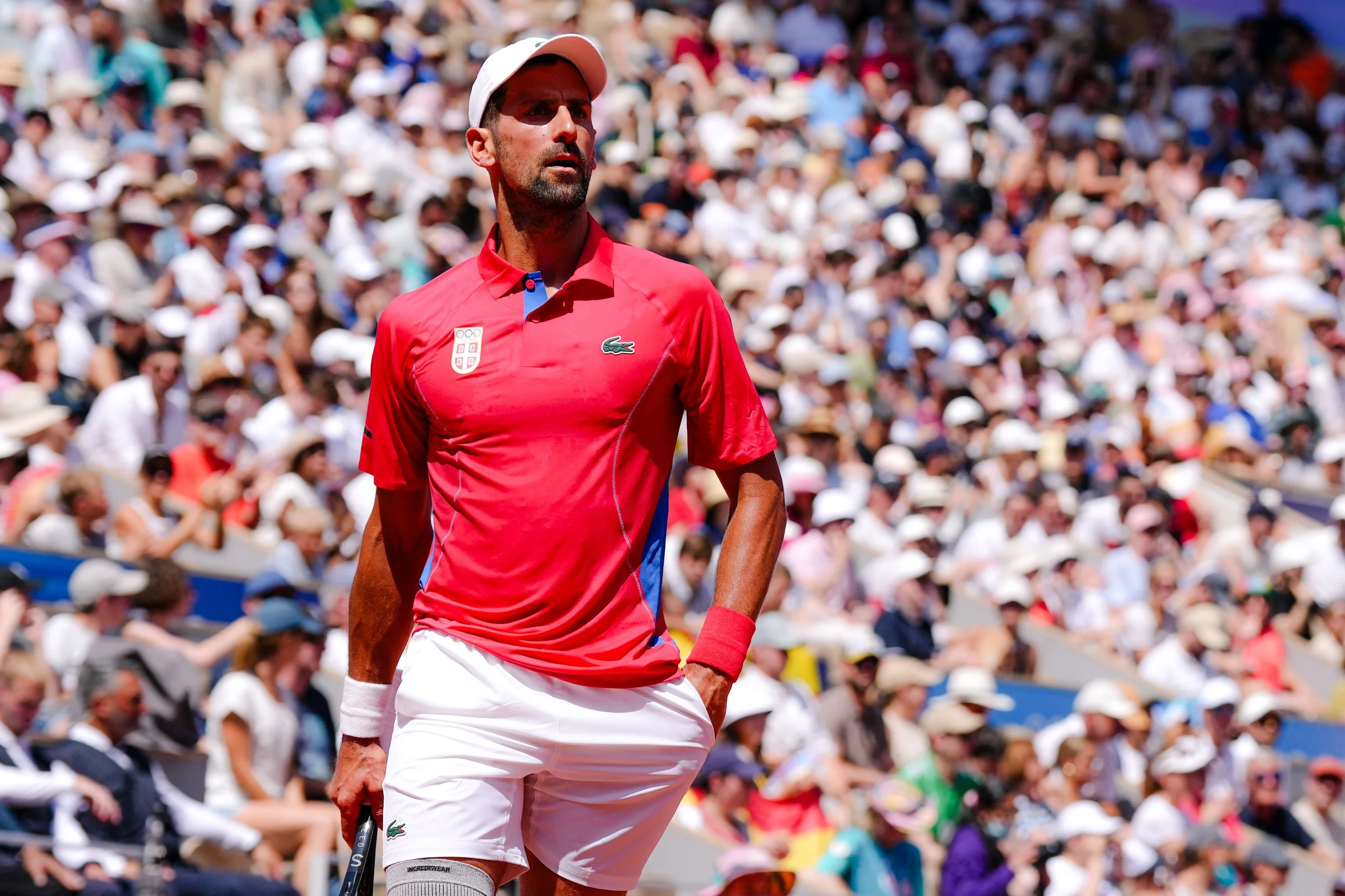 A male tennis player dressed in a red shirt, white shorts, and a white hat, standing on a tennis court during a match, with a large crowd of spectators in the background.